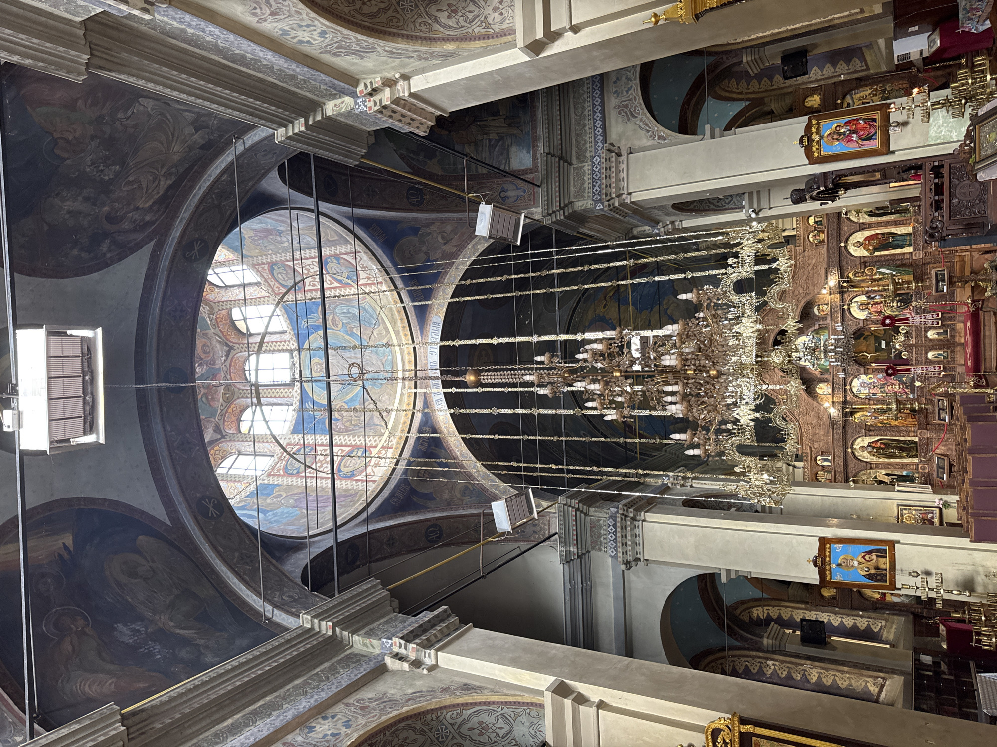 The image shows the interior of a grand, ornately decorated Orthodox church. The perspective is looking upwards towards the ceiling and the altar area. The ceiling features intricate frescoes depicting religious scenes and patterns. A large, circular stained-glass window is visible, allowing natural light to filter in. Several ornate chandeliers hang from the ceiling, illuminating the space.
The altar area, visible on the right side of the image, is adorned with religious icons and carved wooden structures, typical of Orthodox churches. A notable icon depicts figures dressed in red and gold robes, crowned, suggesting a depiction of Jesus and Mary or other significant religious figures.
The overall impression is one of solemnity and grandeur, characteristic of a place of worship. The lighting suggests it is daytime, with natural light coming from the stained-glass window and artificial light from the chandeliers. The architectural style and religious iconography point to a location with a strong Orthodox Christian tradition, consistent with the provided location context of Yambol, Bulgaria. There is no discernible text in the image.