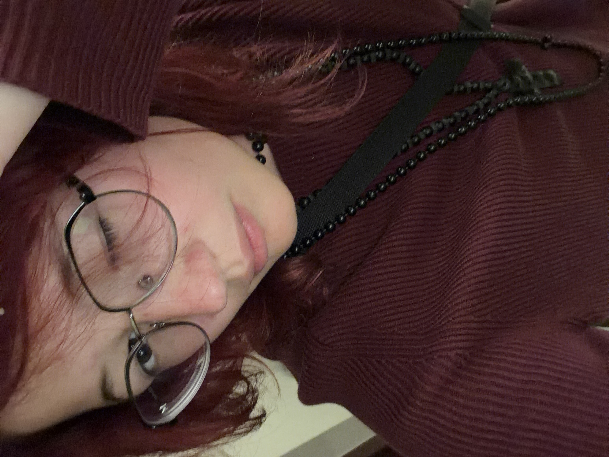 The image is a close-up, selfie-style photograph of a person with red hair wearing glasses and a burgundy ribbed sweater. The person is also wearing a black beaded choker necklace and a black strap across their chest, with another black beaded necklace hanging down, featuring a cross pendant. The background appears to be indoors, with a wall visible on the left side. There is no information or visual cue in the image that would indicate the location is in Chelyabinsk, Russia.
