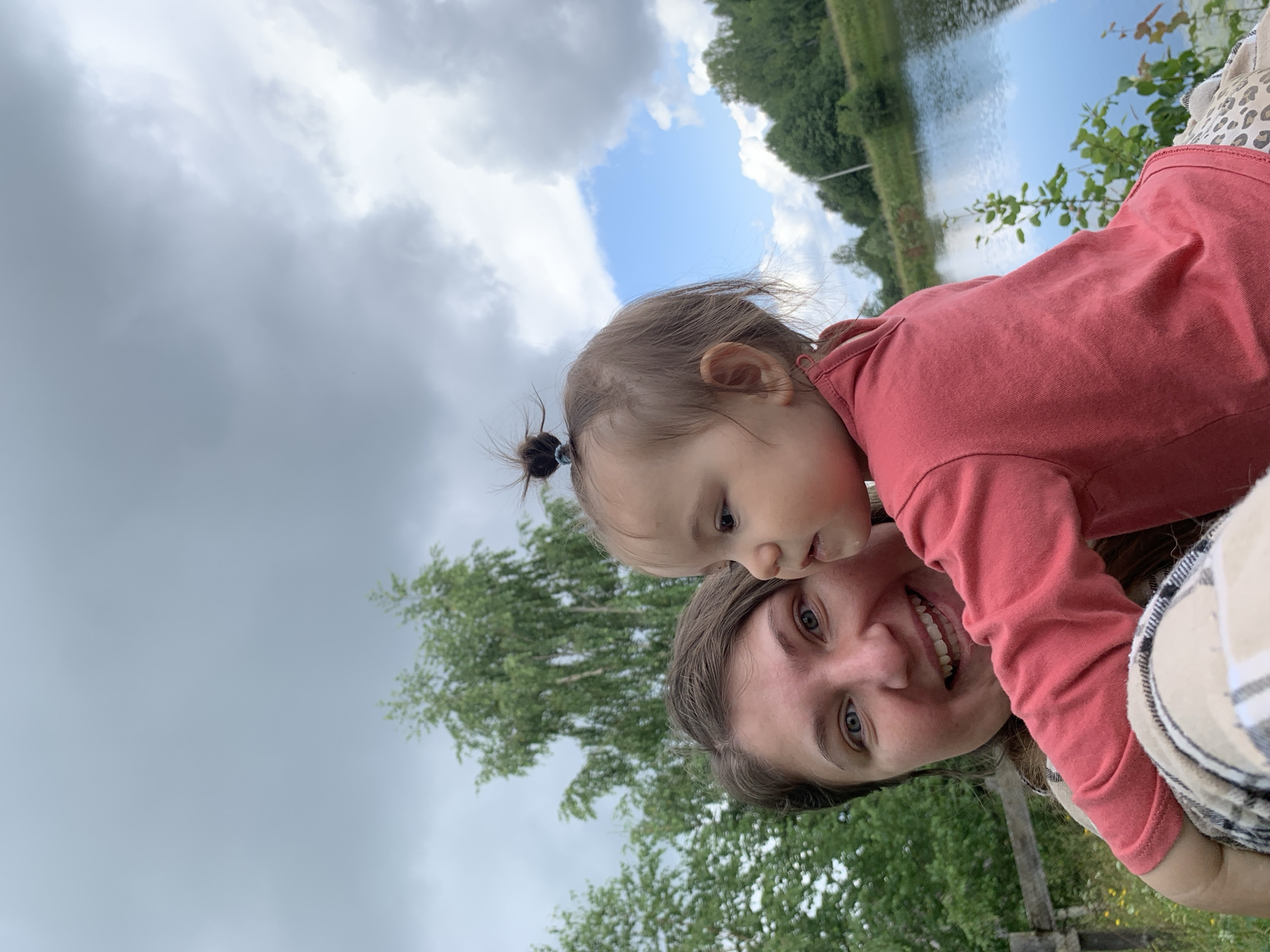 In Zasechnyi, Russia, a selfie is taken outdoors near a body of water. A woman with brown hair and blue eyes smiles at the camera, holding a young child who is looking away from the camera. The woman is wearing a plaid scarf around her neck. The child is wearing a coral-colored shirt and patterned pants. Behind them, a lake is visible, surrounded by trees and a cloudy sky. A wooden fence is present in the foreground.