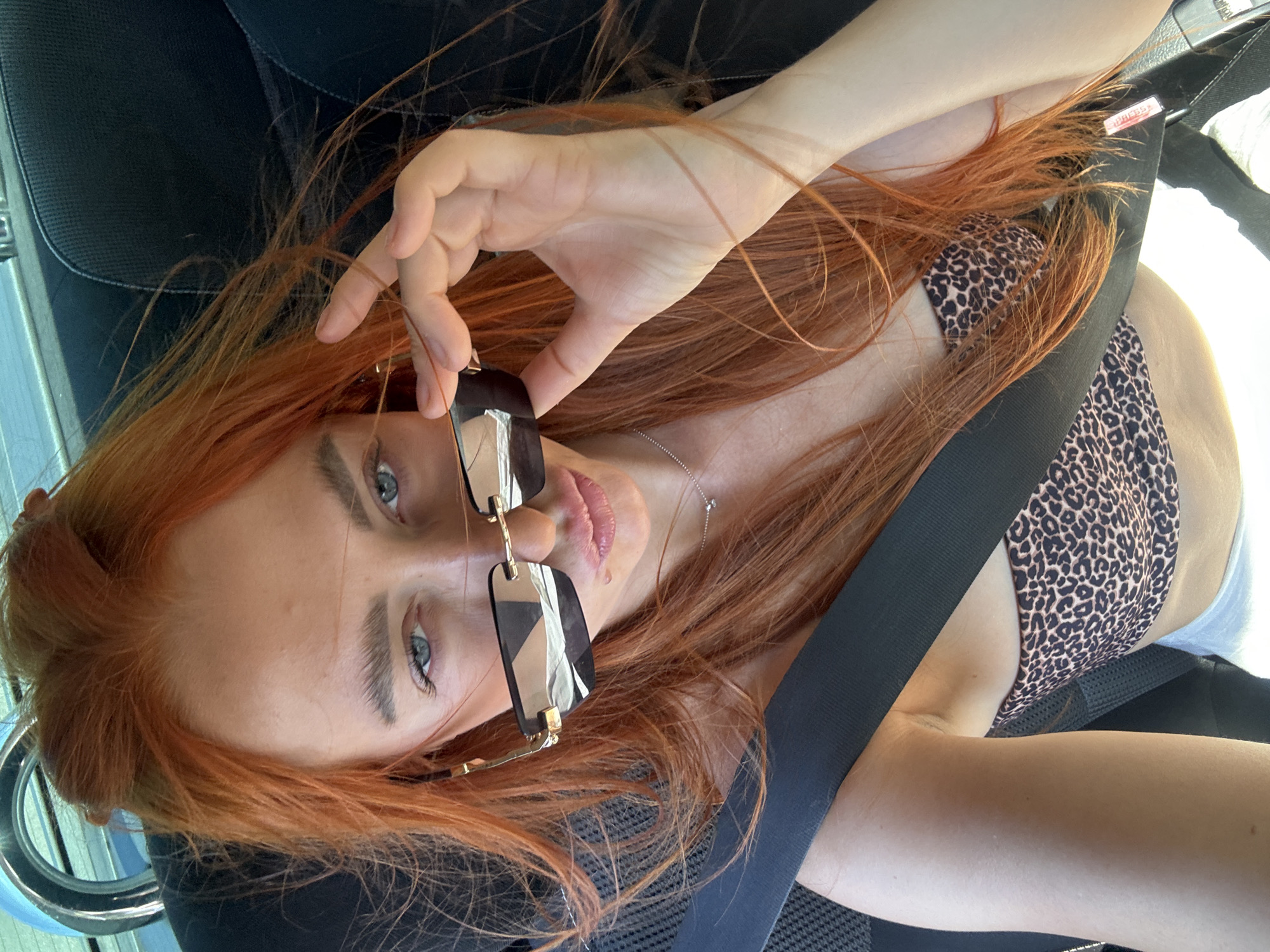 A close-up, selfie-style photograph shows a woman with bright red hair sitting in the passenger seat of a car. She is wearing a leopard-print bikini top and white shorts. Her hand is raised, adjusting a pair of dark-rimmed sunglasses. The woman is looking directly at the camera with blue eyes. The interior of the car is visible, including a black seatbelt across her chest and the headrest. The background is slightly blurred, suggesting movement, and a sliver of what appears to be a road or water is visible through the car window. The scene is consistent with being in Mišići, Montenegro, a coastal town known for its scenic drives.