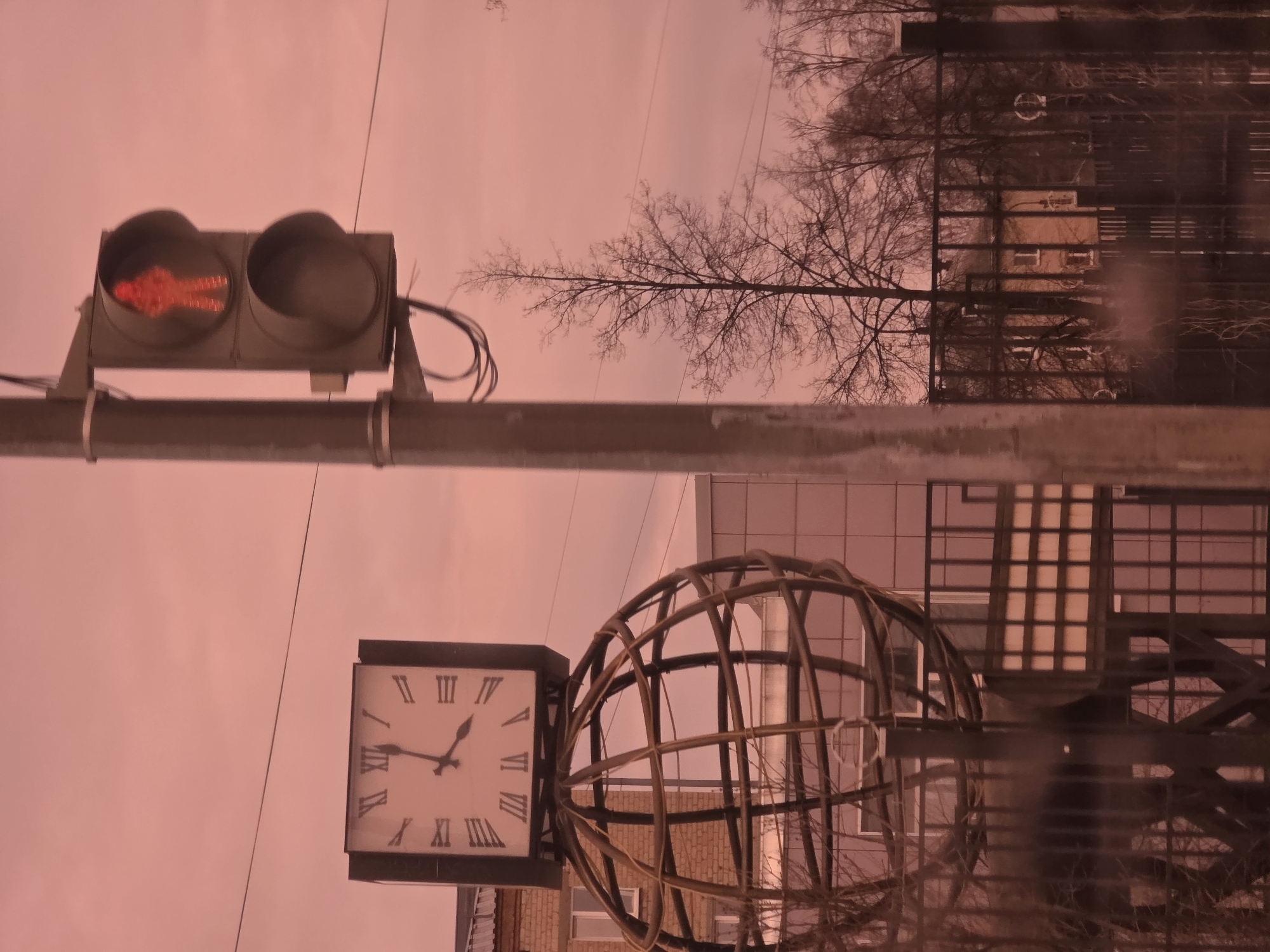 The image shows a street scene in Chaikovsky, Russia, on a day with a pinkish-grey sky. A horizontal pole supports a traffic light displaying a red "pedestrian crossing" signal. Below the light, a large, square clock with Roman numerals is mounted. To the right of the clock, a spherical structure made of interwoven branches or rods adds an artistic element. Behind these structures, buildings with reflective windows and bare trees are visible. The scene appears to be either a reflection or a photograph taken from a low angle, looking upwards.