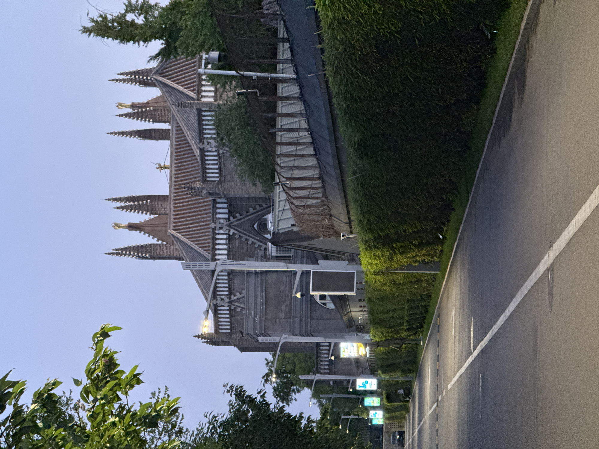 The image shows a street scene in Xicheng District, China, featuring a building with ornate spires reminiscent of European Gothic architecture. The building is constructed of gray stone and features a red-tiled roof. The street runs alongside the building, marked by a white line, and is lit by streetlights. Dense hedges line the edge of the street opposite the building. Street signs are visible in a row along the road. Green foliage, potentially trees, are located in the foreground. The sky is a muted blue-gray, indicating twilight or early evening.