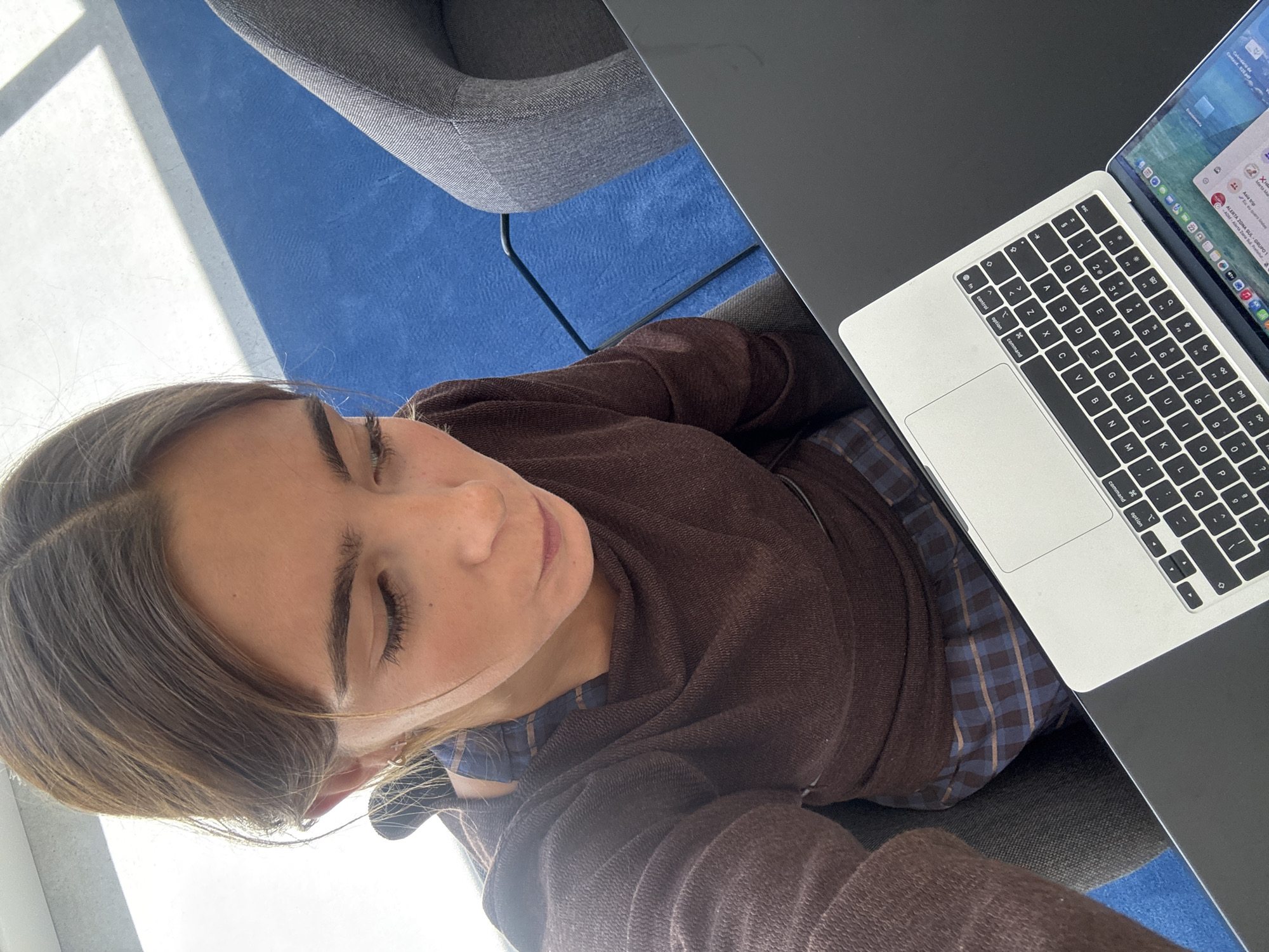 A woman is sitting at a desk in Torres Vedras, Portugal, working on a laptop. The background includes a blue carpet and a grey chair. The woman wears a brown sweatshirt and a blue and brown plaid shirt.