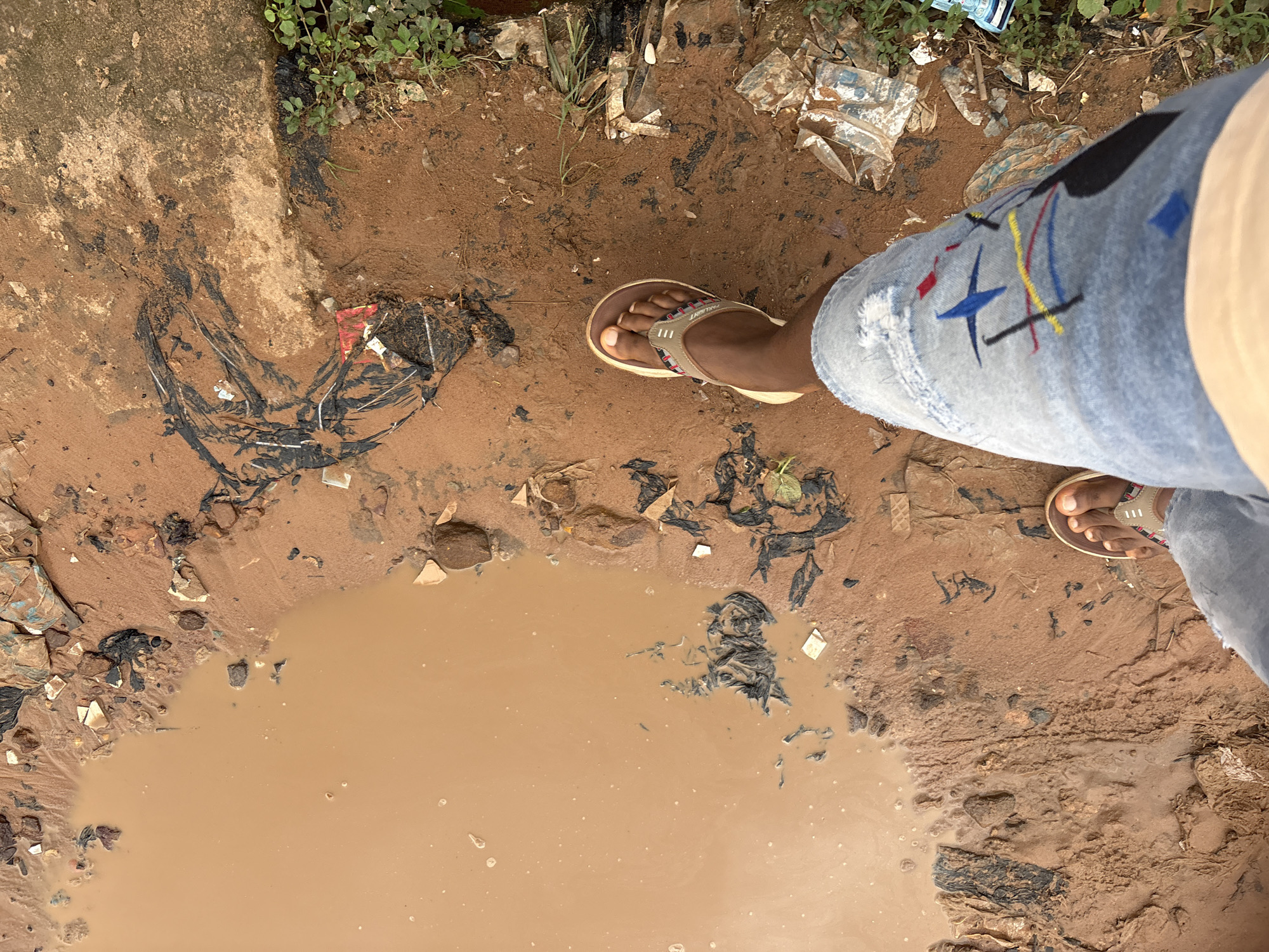 The image shows a person's legs and feet standing on a muddy, debris-strewn path. The ground is wet and appears to be a mixture of reddish-brown mud, sand, and various types of garbage including plastic bags, torn paper, and indeterminate objects. The water forms a large puddle in the upper left portion of the image. The person is wearing light blue jeans with distressed details and colorful embroidery, and a pair of brown and beige flip-flops. The surrounding environment appears to be an outdoor area, possibly a roadside or a neglected urban space. The location is identified as Nkpor, Nigeria, which is known for its commercial activities and sometimes suffers from poor waste management and drainage, leading to such scenes after rainfall.