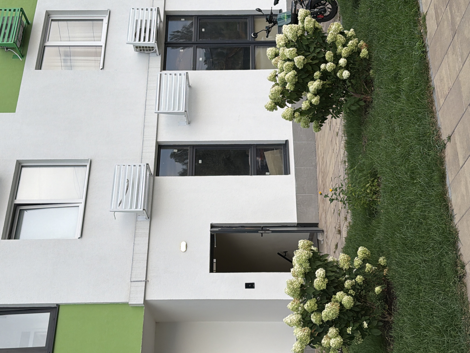 The image shows the exterior of a modern residential building with a light-colored facade, likely in Kyiv, Ukraine. The building features several windows with dark frames, and some sections of the facade are painted bright green. Multiple outdoor air conditioning units are mounted on the exterior walls. In the foreground, there is a patch of green grass and a paved walkway. Two large bushes with white, hydrangea-like flowers are prominent in the scene, and a small yellow flower grows between them. To the right of the image, a black electric scooter or motorcycle is parked. An open doorway reveals stairs leading down inside the building, and a black call box is mounted on the wall next to it. The lighting suggests it is daytime, with some shadows indicating the sun's position.
