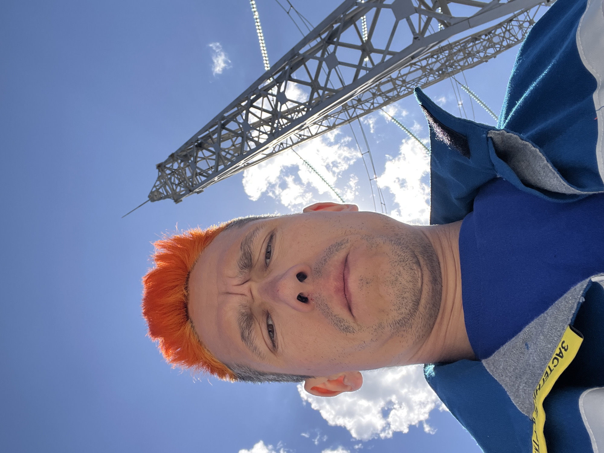 A low-angle selfie depicts a person with orange hair standing under an electrical transmission tower in Chaikovsky, Russia, against a blue sky with scattered clouds. The transmission tower is made of steel and has a lattice structure. The person is wearing a blue shirt.