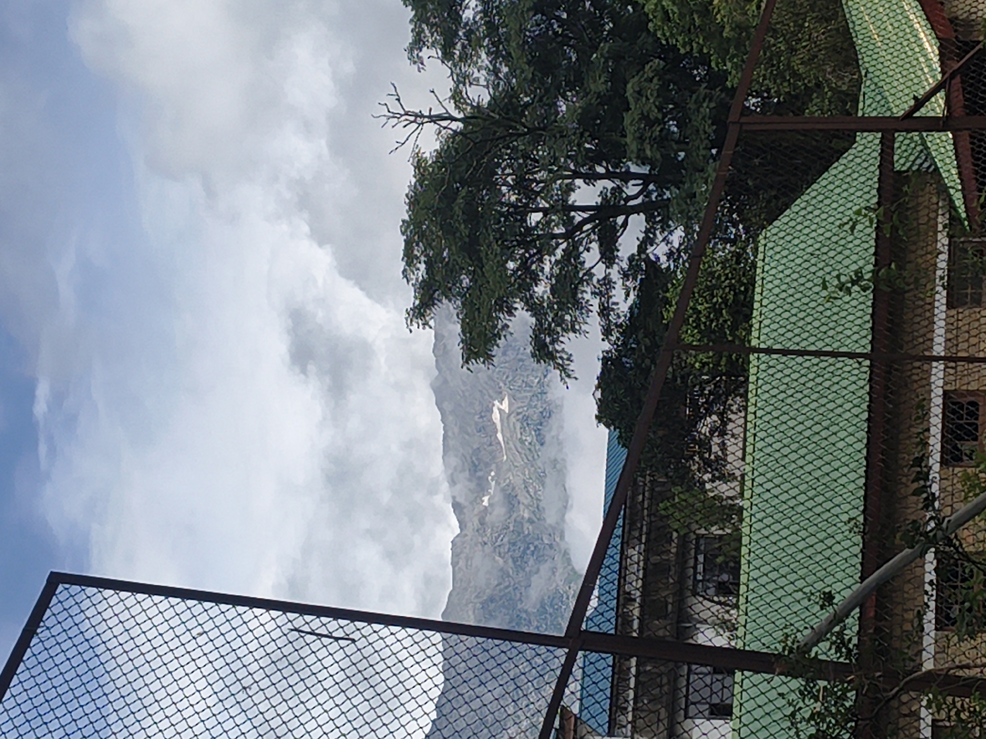 The image captures a view of the Dhauladhar mountain range from Dharamshala, India, under a cloudy sky. In the foreground, a chain-link fence with a dark metal frame partially obscures a building with a green corrugated roof. The building appears to be a single-story structure with some visible windows. Behind the building, a dense canopy of green trees stands out, with the stark branches of a dead tree visible against the sky. The mountains in the background are rugged and rocky, with patches of snow or white rock formations scattered across their slopes. Wisps of clouds drift around the mountain peaks, adding a sense of depth and atmosphere to the scene. The overall impression is one of a natural landscape viewed from an urban or semi-urban setting.