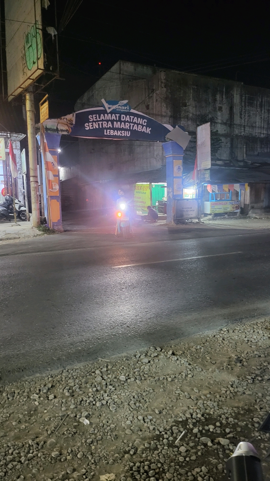 The image shows a street scene at night in Lebaksiu, Indonesia. An archway with the text "SELAMAT DATANG SENTRA MARTABAK LEBAKSIU" is visible across the road. Underneath the archway, a person is standing next to a motorcycle, with its headlight on, illuminating the scene. To the right of the archway, there are food stalls with signs, including one for "GORENGAN TEK TEK". The road is asphalt and appears wet, with gravel on the sides. In the background, there is a large, weathered concrete building. The lighting suggests it is nighttime, with artificial lights from the archway, motorcycle, and stalls contributing to the illumination.