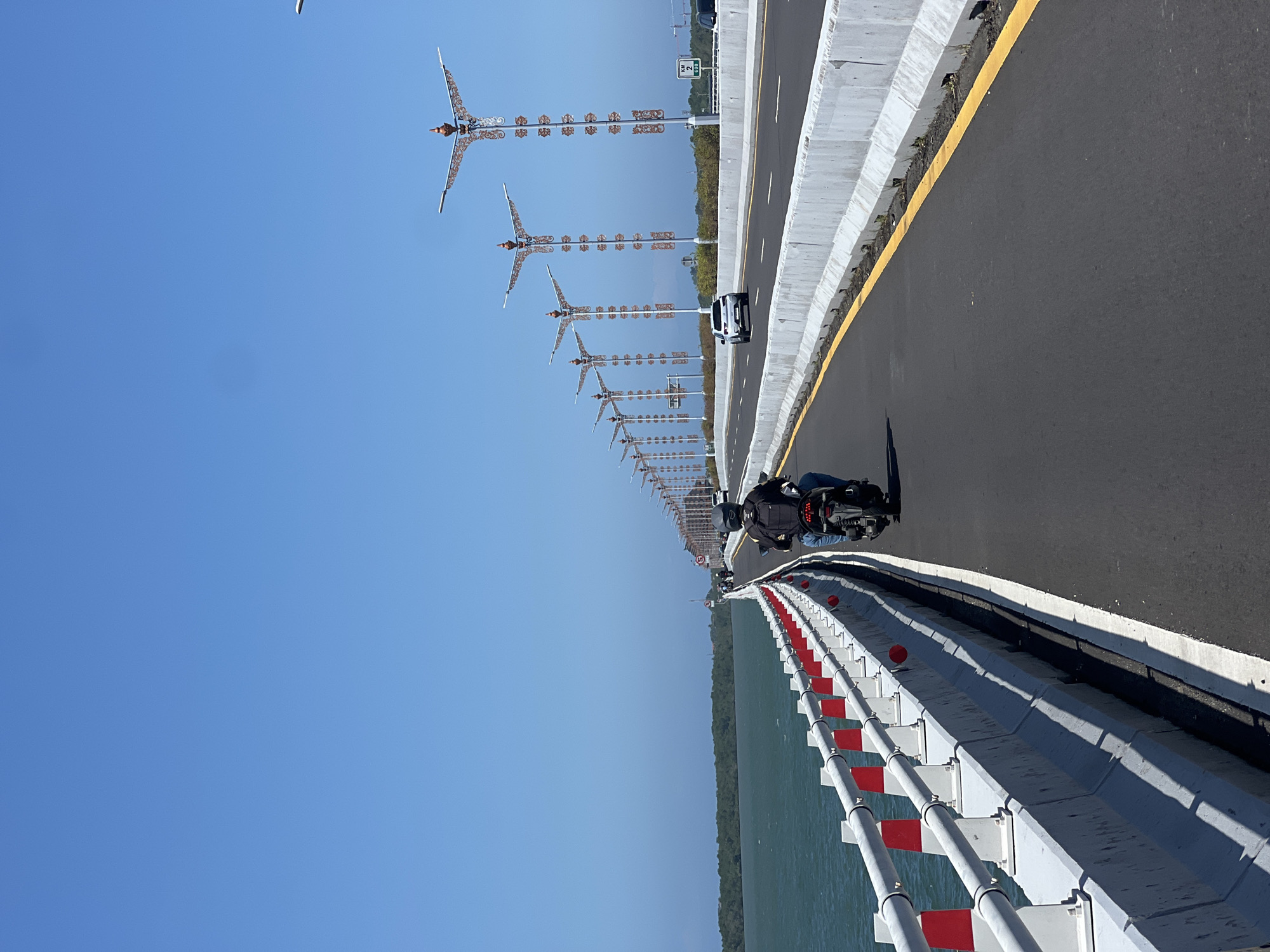 This is a full, eye-level, outdoor shot of a person riding a motorcycle on a wide, paved road on a bridge. The motorcycle is black and the rider is wearing a black helmet and backpack. The road has a yellow line dividing lanes. To the left of the road is a body of water with a green, tree-covered coastline in the distance. To the left of the road is a white and red railing. To the right of the road, there are several decorative lampposts. The sky is clear and blue. A white SUV is visible in the distance on the road. The location is the Bali Mandara Toll Road in Kuta Selatan, Indonesia.