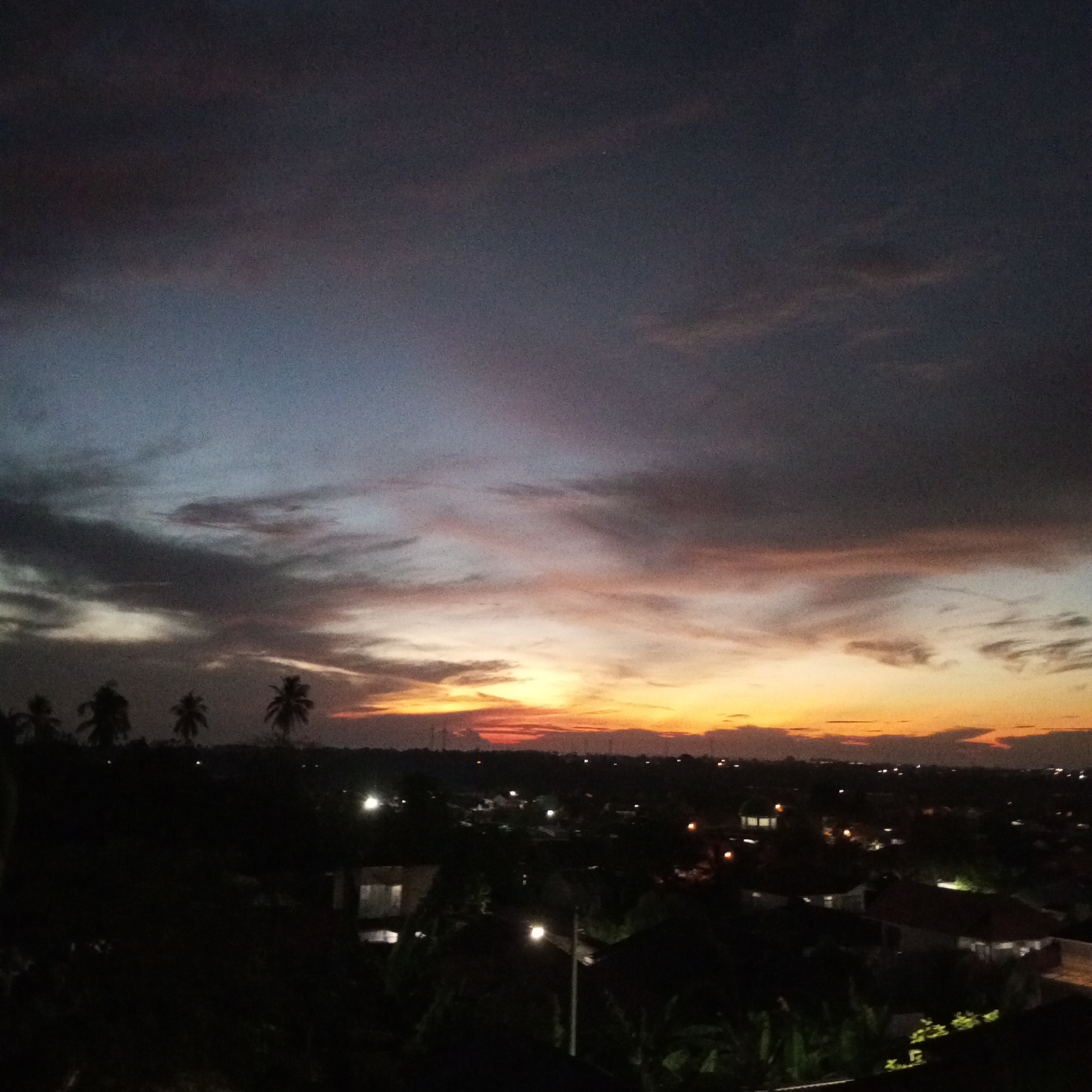 The image depicts a cityscape at dusk in Lhokseumawe, Indonesia. The sky features a dramatic sunset with colors ranging from dark blue and gray to orange and yellow. Several palm trees are silhouetted against the fading light on the left side of the frame. Below the horizon, buildings and streetlights indicate an urban area. The foreground is dark, suggesting the scene is viewed from an elevated position.