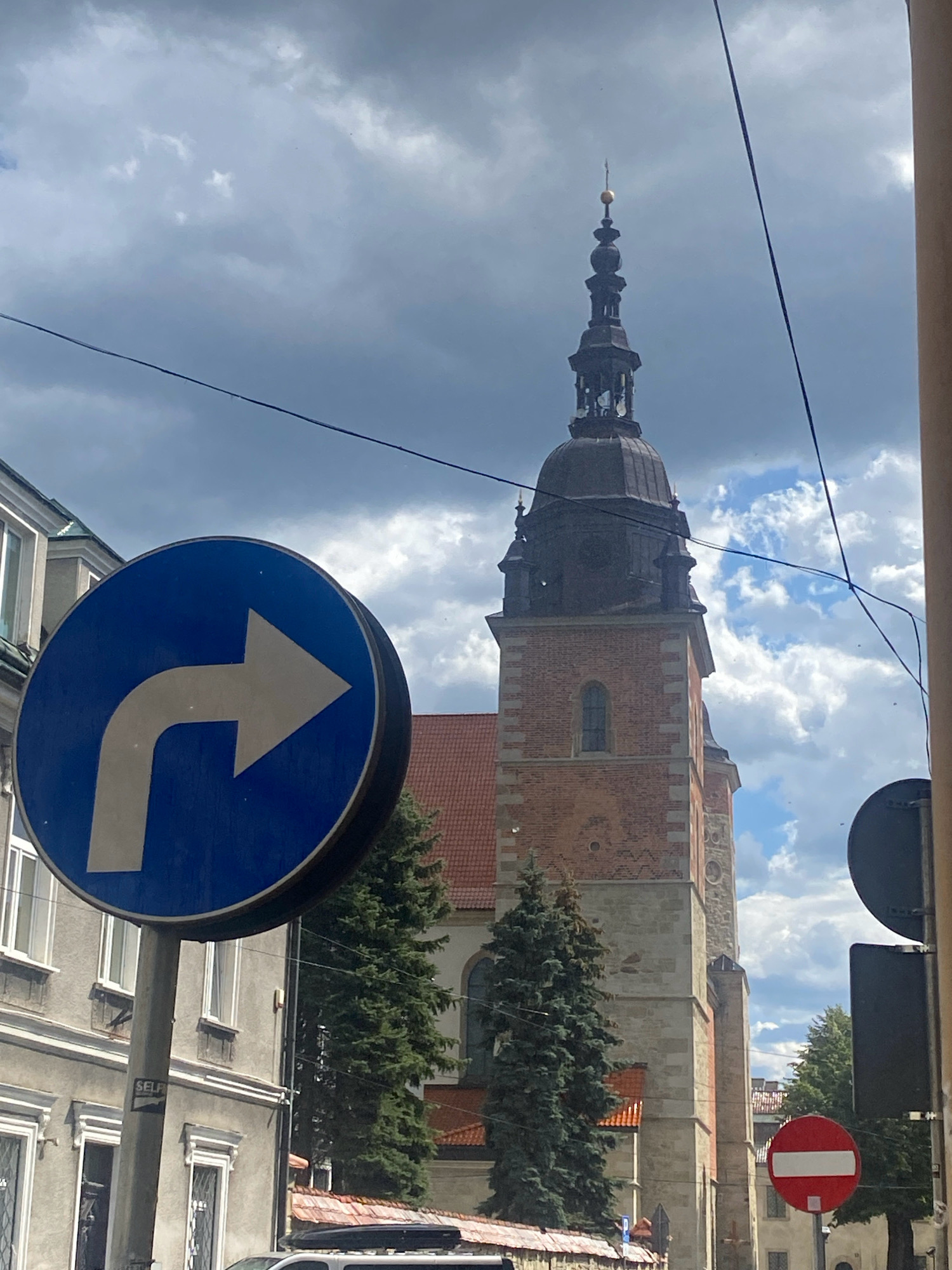 The image shows a street scene in Nowy Sącz, Poland, featuring a prominent church tower. In the foreground, a blue circular traffic sign with a white arrow pointing to the right indicates a mandatory turn. Next to it, a gray building with white-trimmed windows lines the street. Behind these, a tall brick church tower with a dark dome and spire, topped with a golden orb, dominates the skyline. Two evergreen trees flank the church. In the background, more buildings and trees are visible under a sky filled with dramatic, dark clouds interspersed with patches of blue. A red circular "Do Not Enter" sign is also visible in the lower right corner of the frame. There are no people visible in the image, and the scene appears to be captured from a street level perspective.