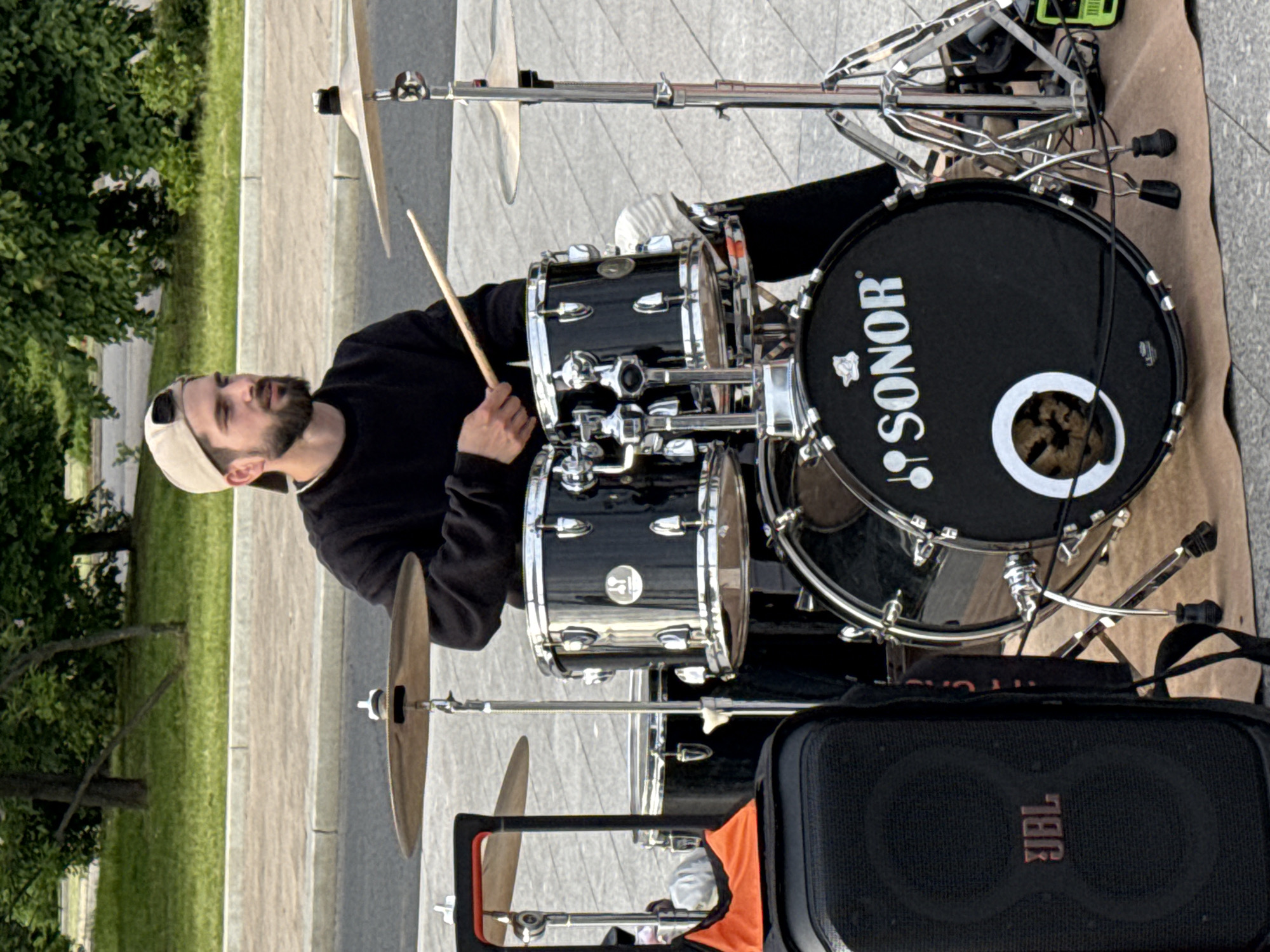 A vertical, eye-level shot captures a male drummer performing outdoors during the daytime. The man, with a beard and wearing a black long-sleeved top and a light-colored baseball cap reversed, is seated behind a black drum kit. He holds drumsticks, appearing focused as he looks slightly upwards, poised to play. The drum kit consists of a bass drum, snare drum, and tom-toms, all in black with chrome hardware, along with cymbals. The bass drum prominently features the white "SONOR" logo and a large white circular cutout. In the lower right foreground, a black JBL speaker is visible, indicating sound amplification. The setting is an urban outdoor space, with a paved ground surface and a background of green grass and leafy trees, suggestive of a park or public square. The natural lighting indicates it is daytime, and the weather appears clear. The overall impression is of a street musician engaged in a performance or practice session. The location context for this scene is Moscow, Russia.
