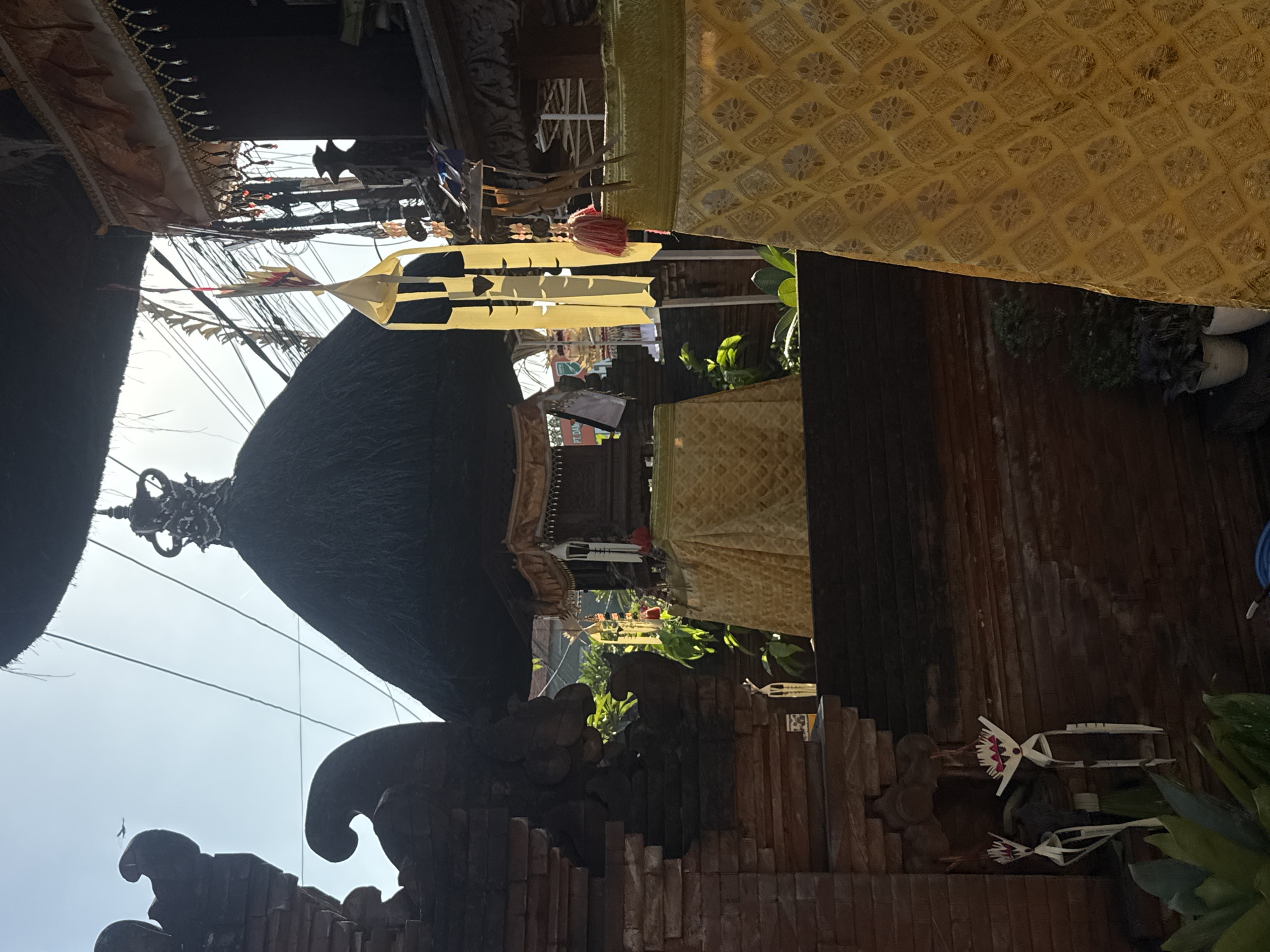 This is a medium shot, taken outdoors, of a traditional Balinese temple complex in North Kuta, Indonesia, bathed in bright, hazy sunlight. The focus is on the architectural elements and ornate decorations, with the sky and surrounding foliage visible in the background.
In the foreground, a richly patterned yellow fabric, likely a ceremonial cloth, drapes over a structure, its intricate diamond motif catching the light. To the left, a weathered brick wall with decorative carved elements frames the scene. Two delicate, straw-like effigies hang from the wall, their red and white heads resembling stylized figures. Lush green plants are visible at the base of the wall.
Dominating the mid-ground is a central shrine with a dark, thatched roof, crowned with an elaborate, dark-colored finial. The shrine is draped with another yellow fabric, this one with a simpler gold-colored pattern. Yellow paper decorations, possibly flags or streamers, hang from the eaves of the structures. Electrical wires crisscross the sky in the upper portion of the image, hinting at the blend of tradition and modernity.
Further back, other temple structures with similar thatched roofs and ornate details are visible, along with more hanging decorations. The overall impression is one of solemnity and cultural richness, with the sunlight casting strong highlights and shadows, adding depth and texture to the scene. There are no people clearly visible in the image.