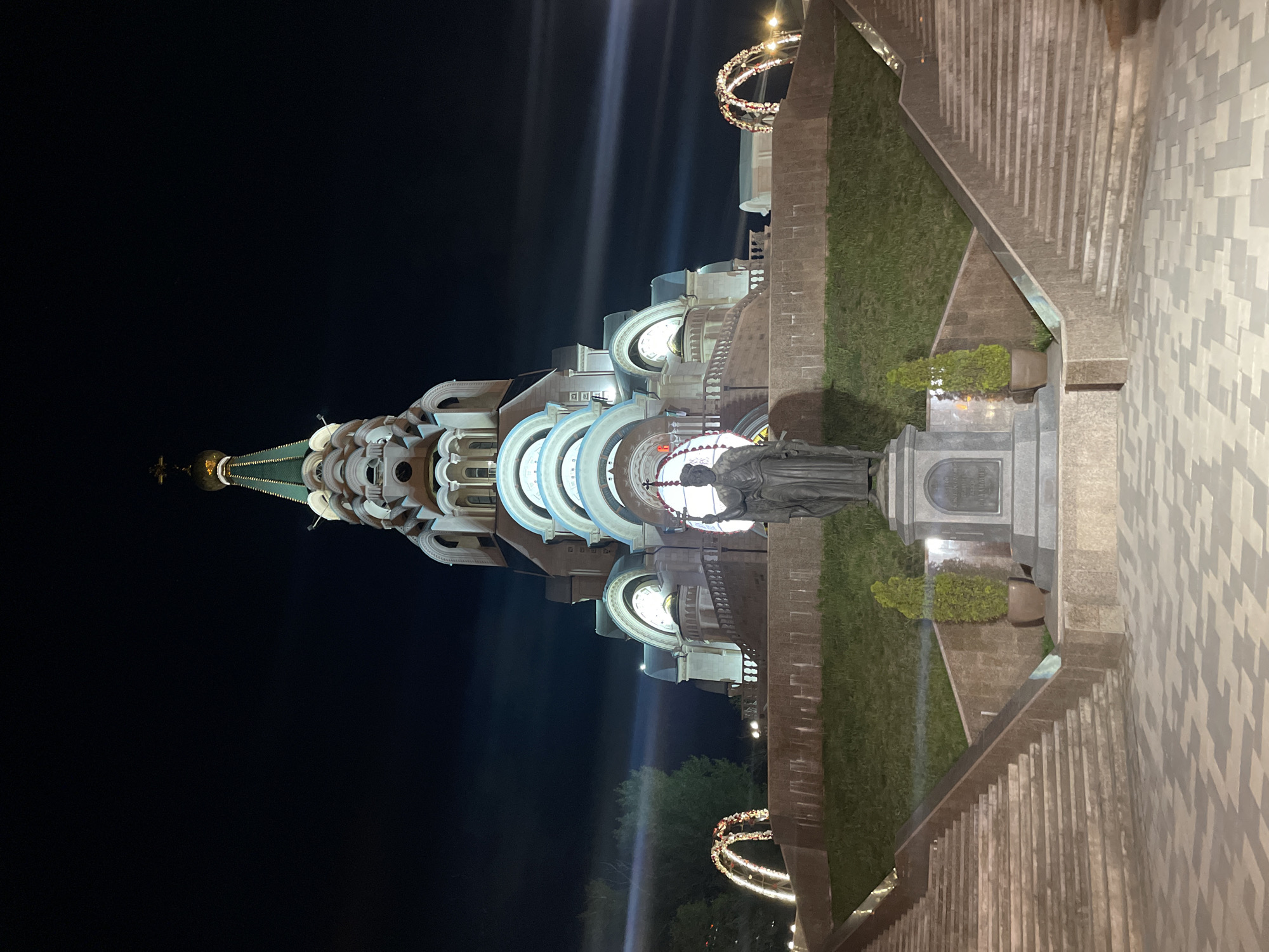 At night, the image captures the Cathedral of St. George the Victorious in Novokuybyshevsk, Russia. The cathedral's architectural details are illuminated, showcasing its spire and arched structures. A statue stands prominently in the foreground, near meticulously landscaped greenery. Stairs with stone banisters lead to the scene. The sky is dark, emphasizing the artificial lighting on the building and surrounding area.