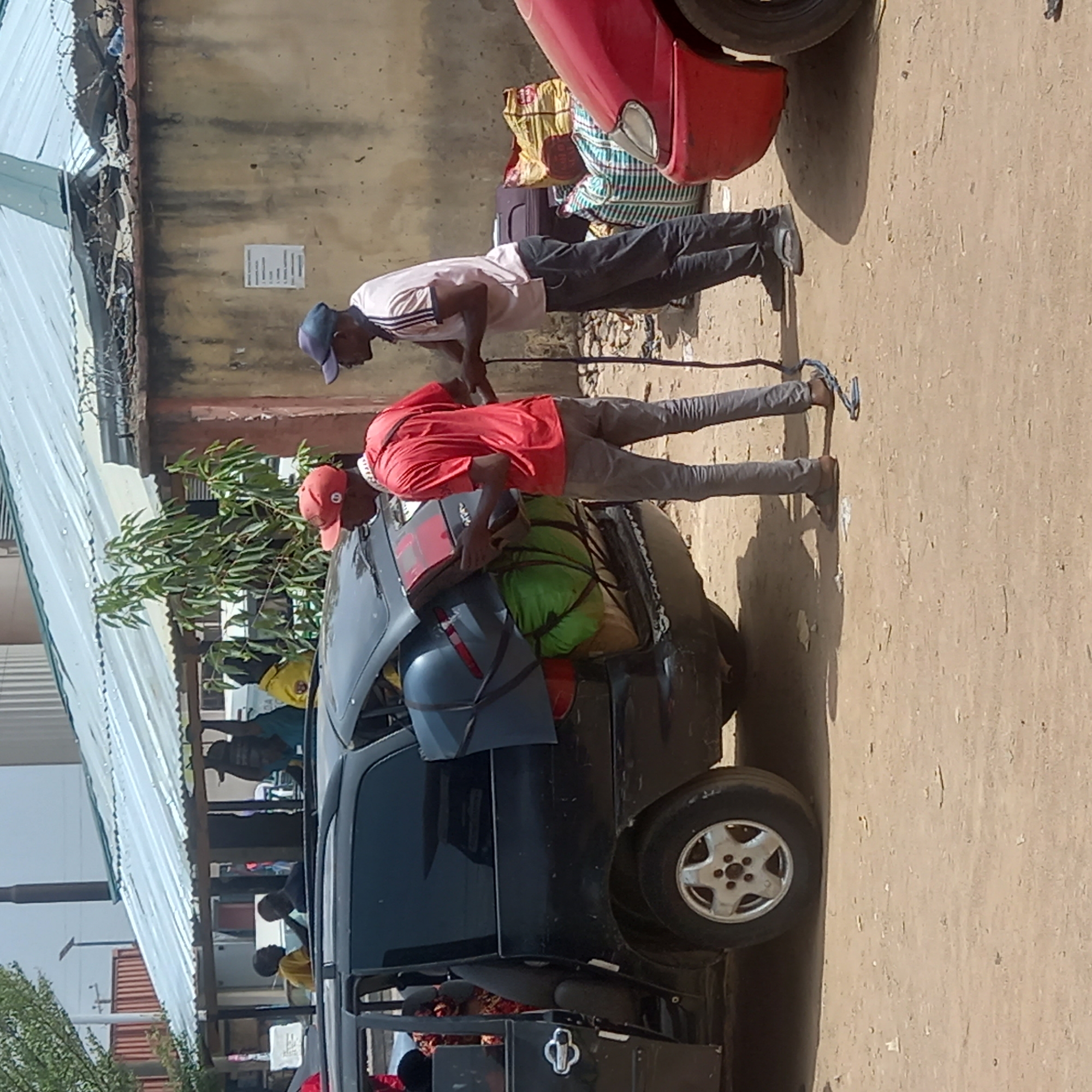 This image captures an outdoor scene in Dadi Riba, Nigeria, under bright daylight, suggesting sunny weather. The central focus is a black car, heavily loaded with an assortment of luggage on its roof. The cargo includes various bags, suitcases, and bundled items in colors like green, yellow, and brown, all secured with ropes or straps.
Two men are prominently featured, actively engaged with the loaded vehicle. On the left, a man wearing a light-colored polo shirt, dark trousers, and a blue baseball cap is leaning forward, possibly assisting with the loading or checking the car's contents. To his right, another man, dressed in a red long-sleeved shirt, light grey trousers, and a red cap, is also leaning over the rear of the car, with his hands positioned on some of the luggage, indicating he is either securing or adjusting the load. A dark blue/grey bag or backpack is visible resting on the back of the car.
In the background, a building with a light brown or yellow wall and a corrugated metallic roof runs along the left side. Barbed wire is visible along the edge of the roof. Green foliage can be seen growing between the building and the car. Further back, beneath the roof structure, other people and simple stalls or structures are discernible, hinting at a bustling market or public area. The ground is a light-colored, unpaved surface, likely dirt or sand. A partial view of a red car is visible in the upper right quadrant of the image.
On the wall behind the men, a white rectangular sign with some small, illegible text is present. The overall activity suggests preparation for travel or the transport of goods, common in local transit points.