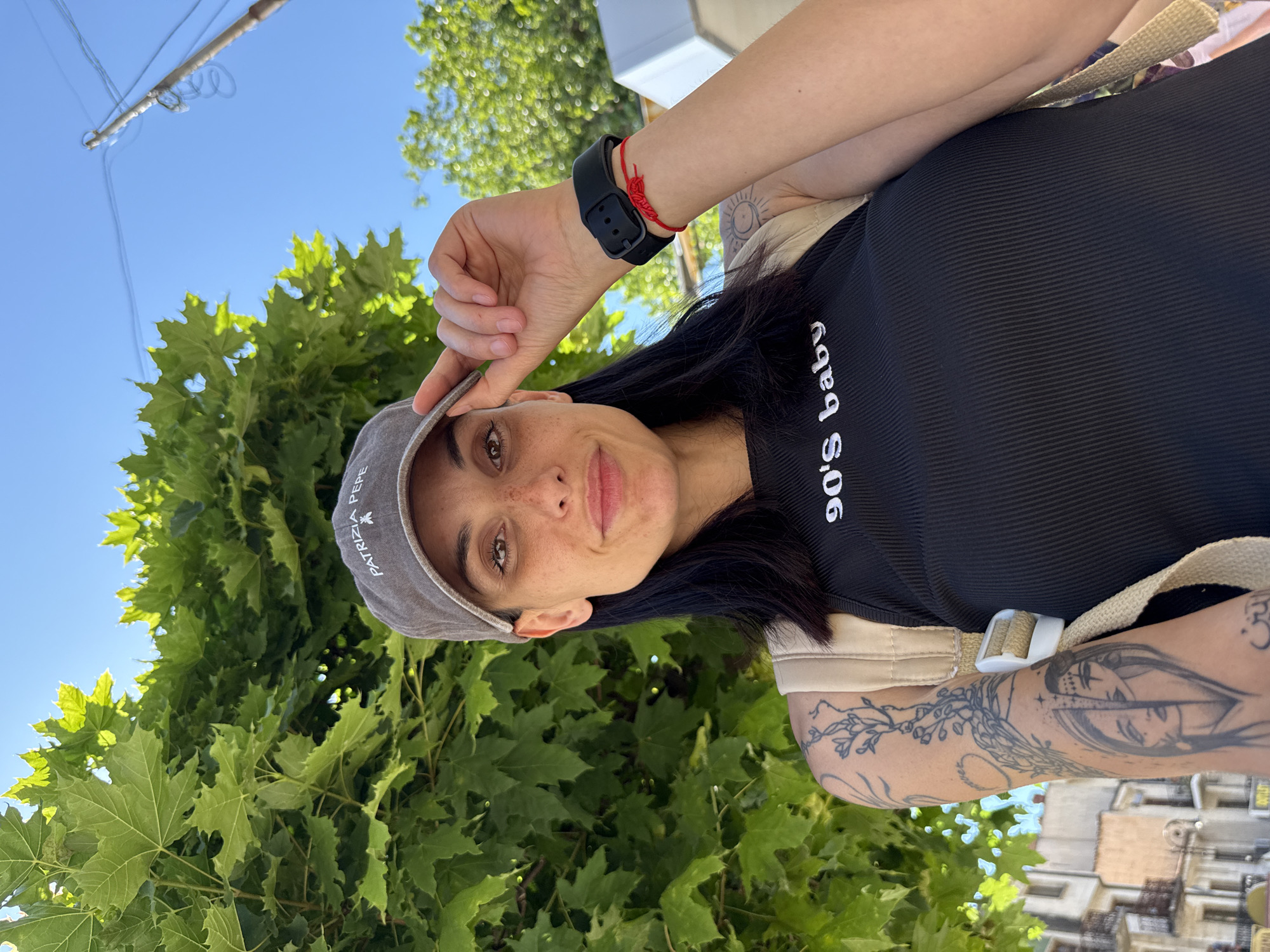 A woman stands outside in Veliko Tarnovo, Bulgaria, positioning the brim of her gray "Patrizia Pepe" baseball cap. She wears a black tank top with "90's baby" printed on it. The woman has dark hair, freckles, and a black smartwatch on her wrist, adorned with a red string bracelet. She has multiple tattoos on her visible arm. She is standing in front of a building and a tree. The sky is blue and visible at the top of the frame.