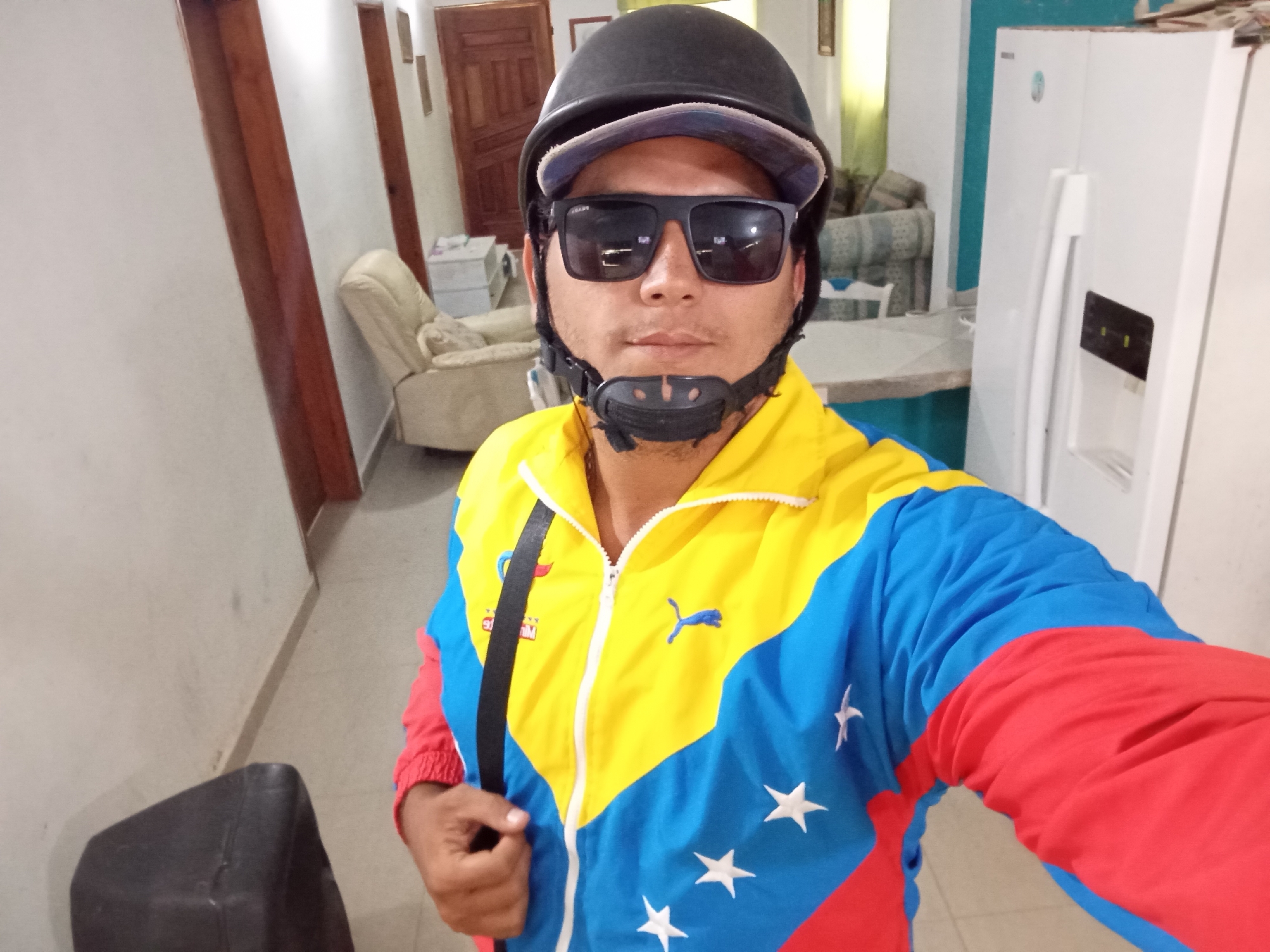 A man wearing a black helmet, sunglasses, and a Venezuela-themed jacket (yellow, blue, and red with white stars) stands indoors, likely in Juan Griego, Venezuela. A black strap crosses his chest, possibly from a bag. Behind him, there's a hallway, furniture, and a white refrigerator. The walls are white.