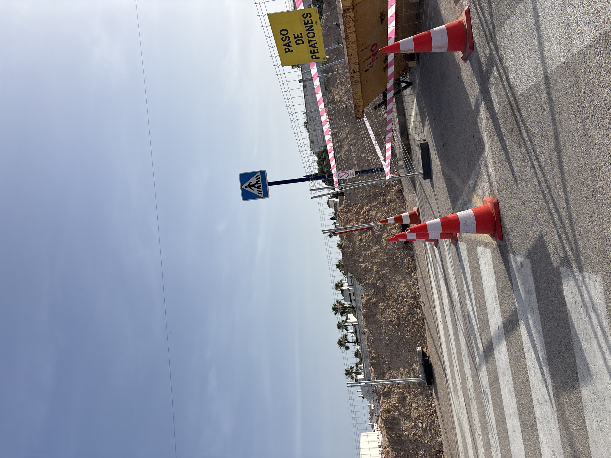 The image depicts a construction site near a pedestrian crossing in Orihuela, Spain. A clear blue sky with a single thin wire stretches across the top. In the foreground, a pedestrian crossing with white stripes is visible on an asphalt road, with long shadows cast across it. Several orange and white traffic cones are placed on the road and sidewalk, delineating the construction area. A temporary metal fence with pink and white striped caution tape runs along the perimeter of the site. Behind the fence, a large pile of dirt and debris is present, with some palm trees and white buildings visible on a ridge in the background. A yellow sign with black text that reads "PASO DE PEATONES" (Pedestrian Crossing) is attached to the fence, indicating the direction of the crossing with an arrow. A blue sign with a white pedestrian symbol is also visible. A large yellow skip is positioned within the fenced-off area, suggesting ongoing construction work. There are no people visible in the image. The overall scene conveys a temporary disruption to normal traffic flow due to construction.