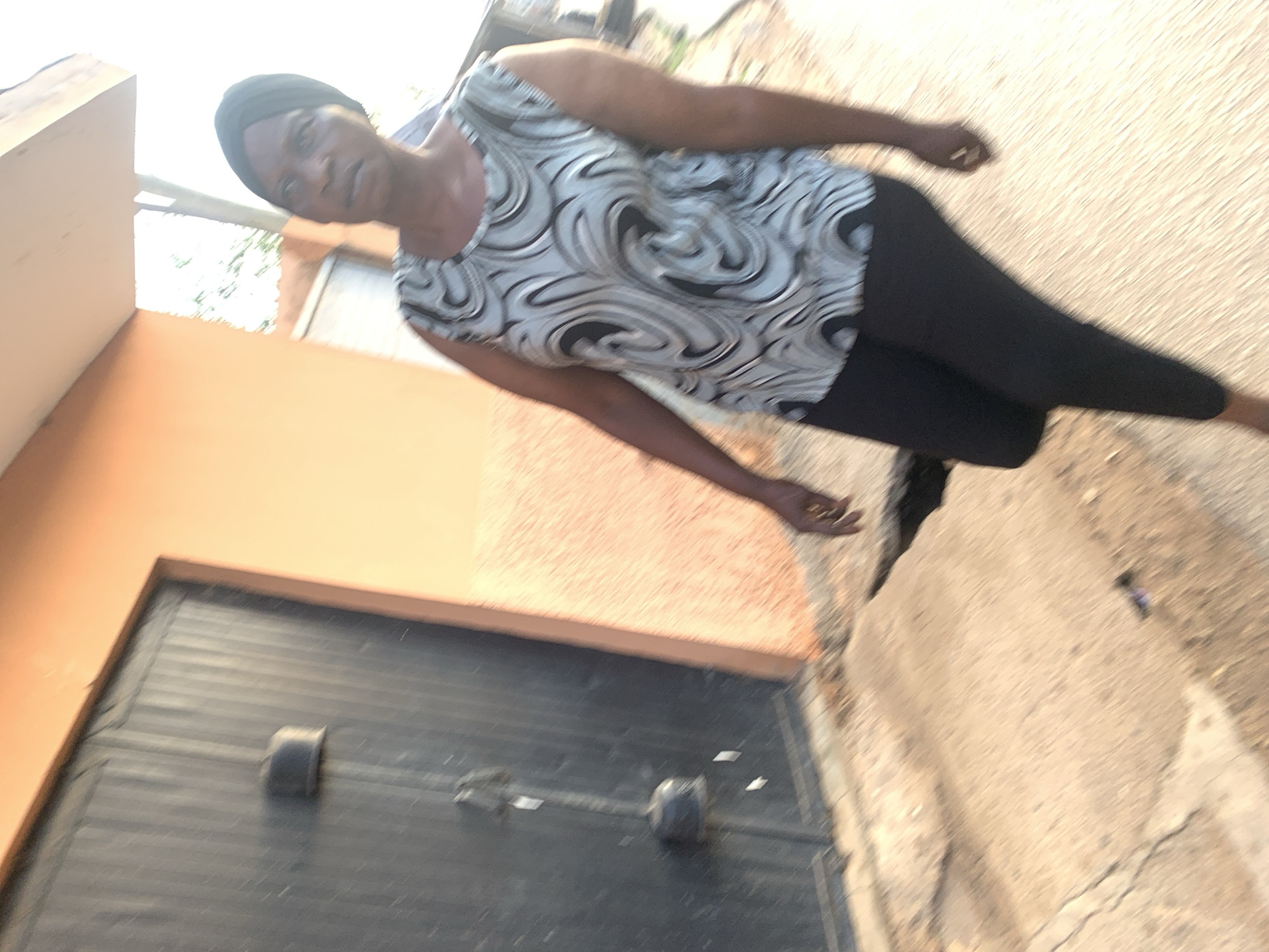 A woman is walking past a dark metal door in Jos, Nigeria. She is wearing a black headwrap and a sleeveless shirt with a swirling grey and white pattern. She is also wearing black leggings. The building appears to be painted a pale orange. The ground in front of the building is concrete and shows signs of disrepair with cracks and a hole. The lighting suggests it is daytime, possibly late afternoon, with a warm glow.