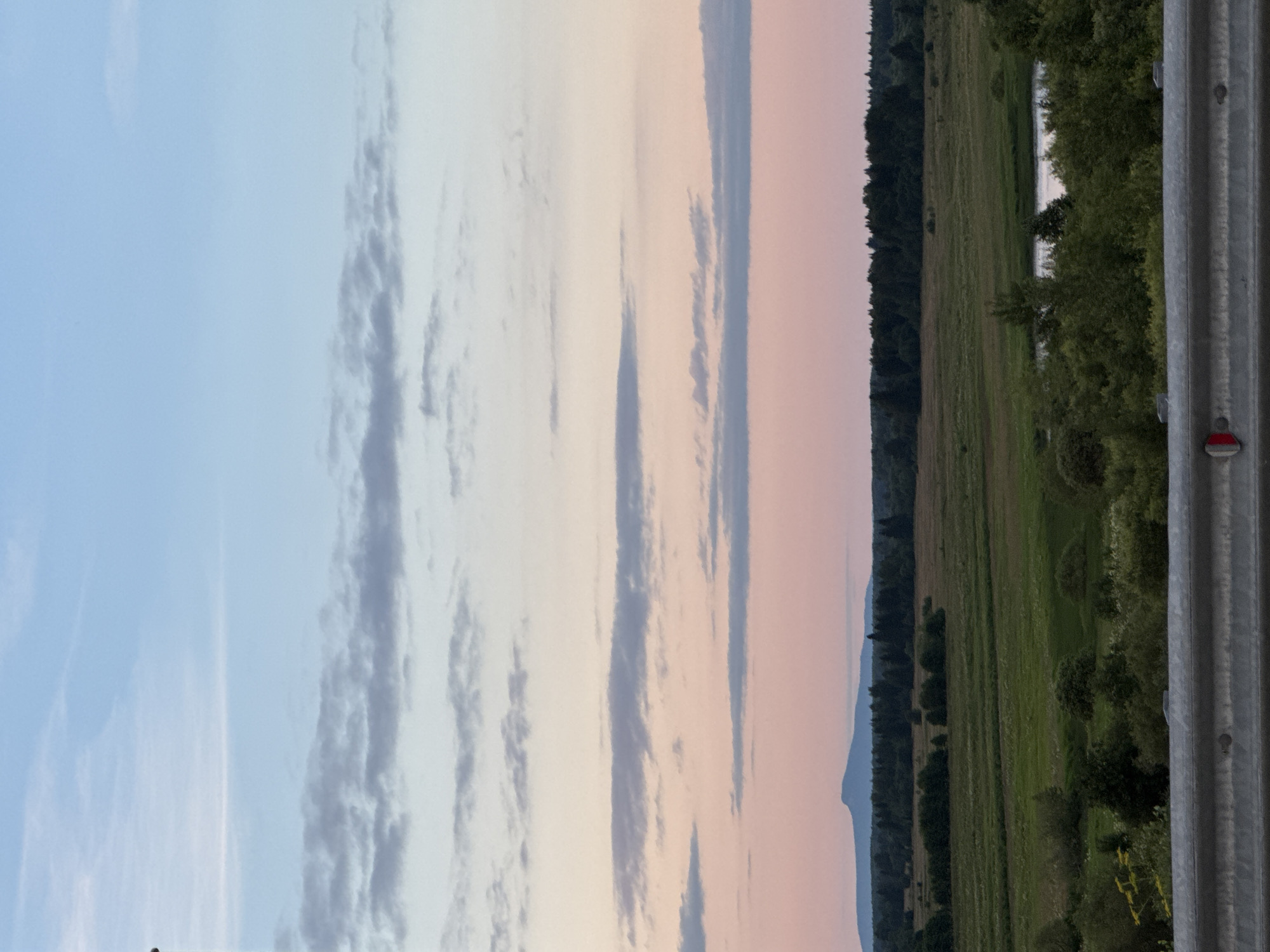 A wide shot captures a pastoral landscape under a twilight sky. The sky transitions from a pale blue at the top to soft pinks and oranges near the horizon, dotted with wispy clouds. Below the sky, a range of rolling green hills and fields stretches across the middle ground, interspersed with dark green forests. A lone mountain peak is visible in the far distance on the left. In the foreground, a metal barrier runs horizontally across the bottom of the image, obscuring the immediate ground. The overall scene is serene and natural, with no signs of human activity or structures other than the foreground barrier. The location is identified as Ryabinino, Russia.