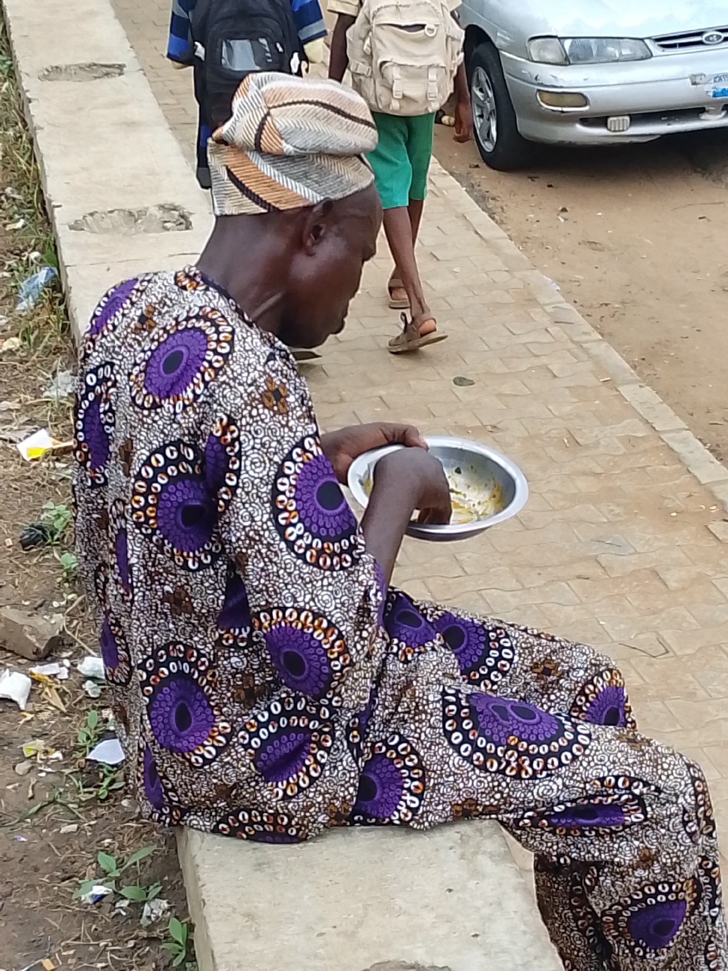 A man in a traditional African outfit with a vibrant purple and white geometric pattern sits on a concrete ledge, eating from a metal bowl. He wears a patterned headscarf that complements his attire. His gaze is directed downward, focused on his meal. Beside him, other individuals are visible, including a person in a backpack and green shorts walking away from the camera, and a silver car parked nearby on a paved surface. The ground is a mix of dirt and paving stones, with some scattered litter visible. The overall scene suggests an outdoor setting, possibly a roadside or marketplace. The lighting indicates it is daytime.