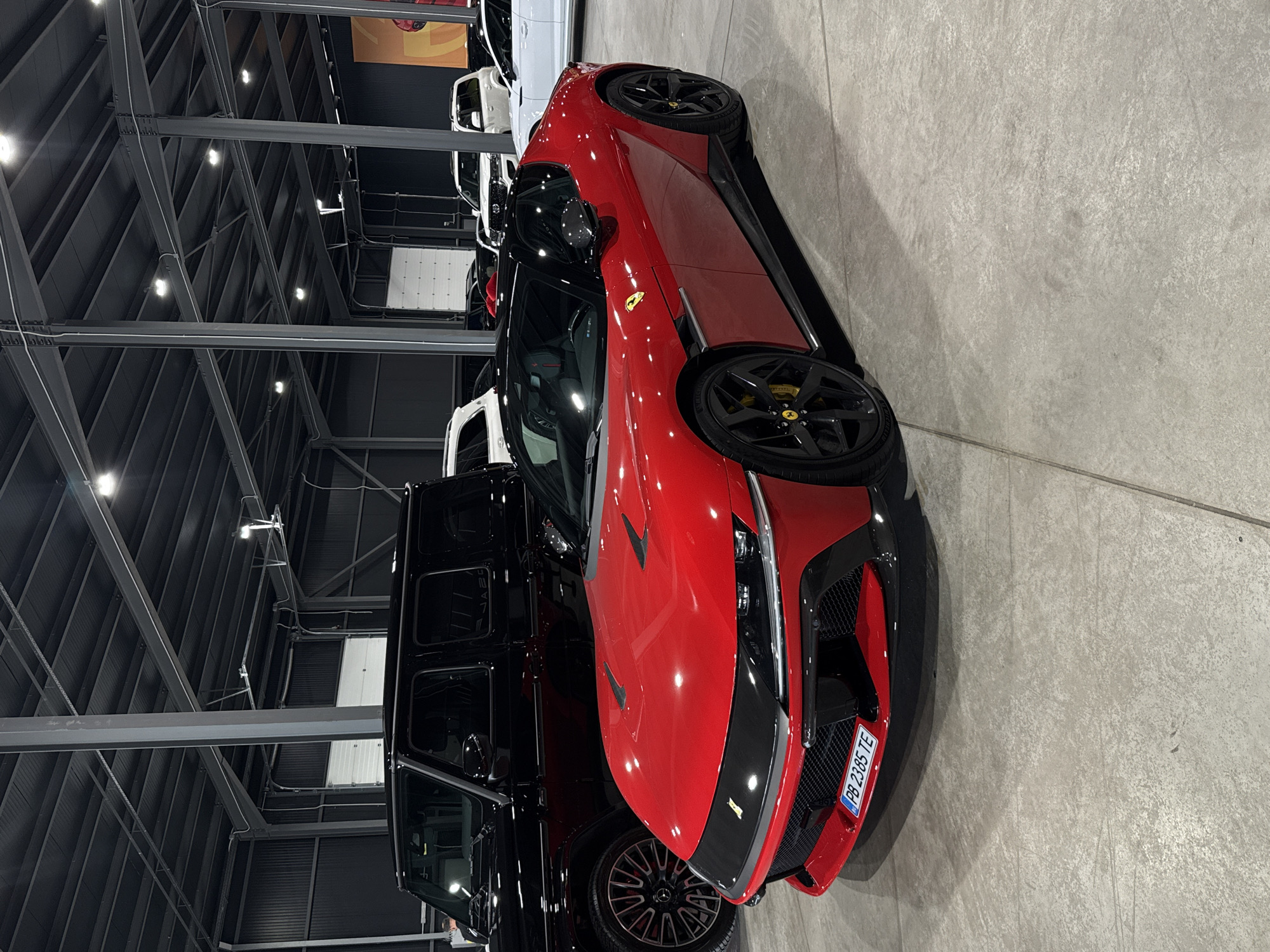 A red Ferrari is parked in a showroom alongside a black Mercedes-Benz G-Class. Other vehicles are visible in the background. The license plate on the Ferrari reads "PB 2385 TE." The showroom is likely in Plovdiv, Bulgaria, given the license plate format, which is used in the Plovdiv province. The showroom's interior features a high, exposed ceiling with metal beams and lighting fixtures.