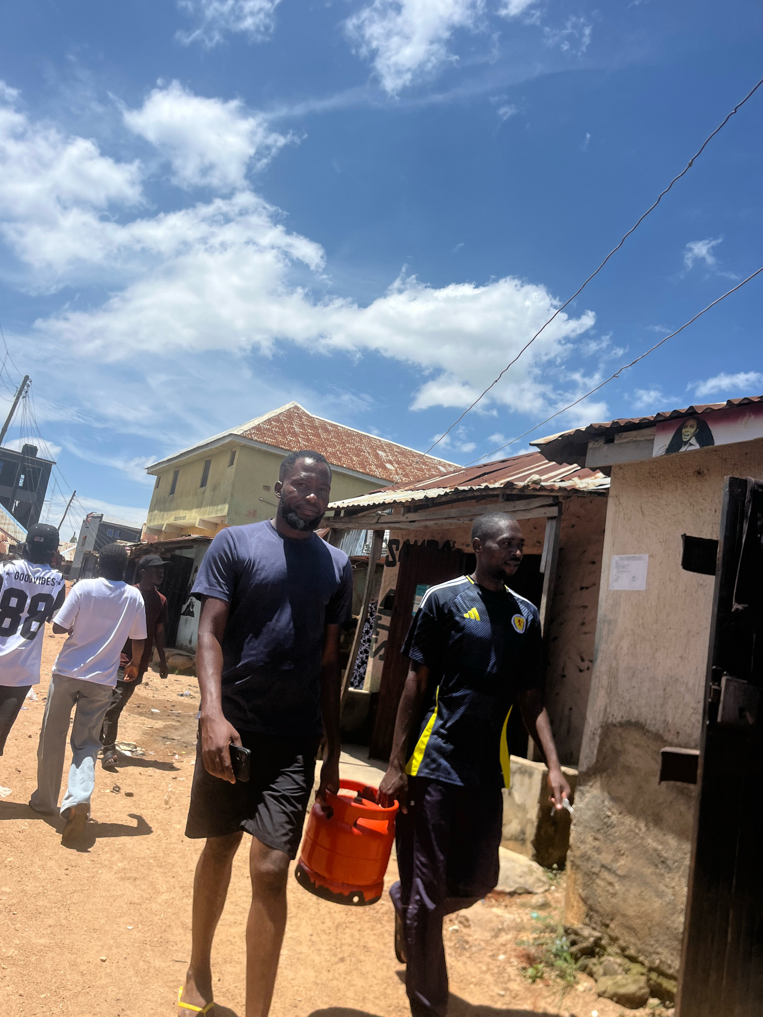 In the city of Jos, Nigeria, two men walk along a dirt road under a partly cloudy blue sky. The man on the left, wearing a dark blue t-shirt and black shorts, carries a bright orange gas cylinder. The man on the right, in a dark blue and yellow striped shirt and dark shorts, holds a cell phone in his left hand. Behind them, other individuals are visible walking away from the camera. The background features a mixture of buildings with corrugated iron roofs and some with yellow painted walls, typical of Jos urban areas. Power lines are strung overhead. The ground is covered in reddish-brown dirt.