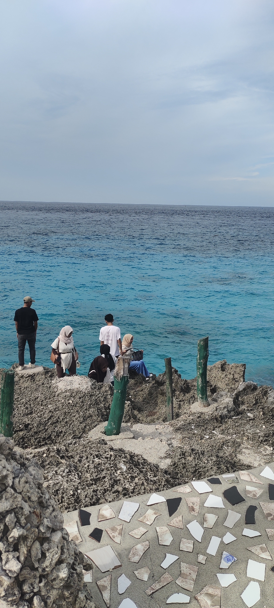 In Sabang, Indonesia, a group of six individuals, some wearing hijabs, are gathered on a rocky shore overlooking the ocean. Two men are standing on the left side of the frame, facing the water. One man is wearing a black t-shirt and a cap, while the other is in a white t-shirt. Three women are positioned on the rocks in the center and right of the frame, two of whom are wearing hijabs. One woman is seated and looking at her phone. Another individual, partially obscured, is crouched on the rocks. The ocean displays a vivid turquoise hue, transitioning to a darker blue towards the horizon, under a partly cloudy sky. Several weathered wooden posts, some painted green, are scattered among the rocks. The foreground features a mosaic of broken ceramic tiles embedded in concrete.