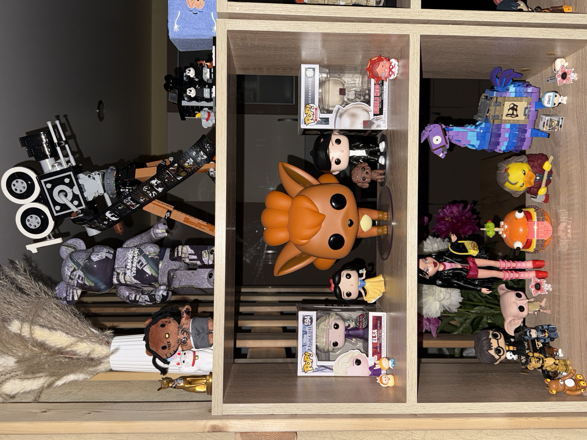 The image displays a collection of pop culture figurines arranged on wooden shelves, likely in a personal collection. The shelves are filled with various Funko Pop figures, including characters from Disney's Frozen (Elsa, Snow White), a Pokémon (Vulpix), and Silent Hill (Bubble Head Nurse). There are also other collectible items such as a Bearbrick figure, a Lego movie camera, and a Lego Llama from Fortnite. The overall setting appears to be an indoor display shelf. The image does not contain any information or visual cues that would allow for the determination of its location in the city of Khimki, Russia.