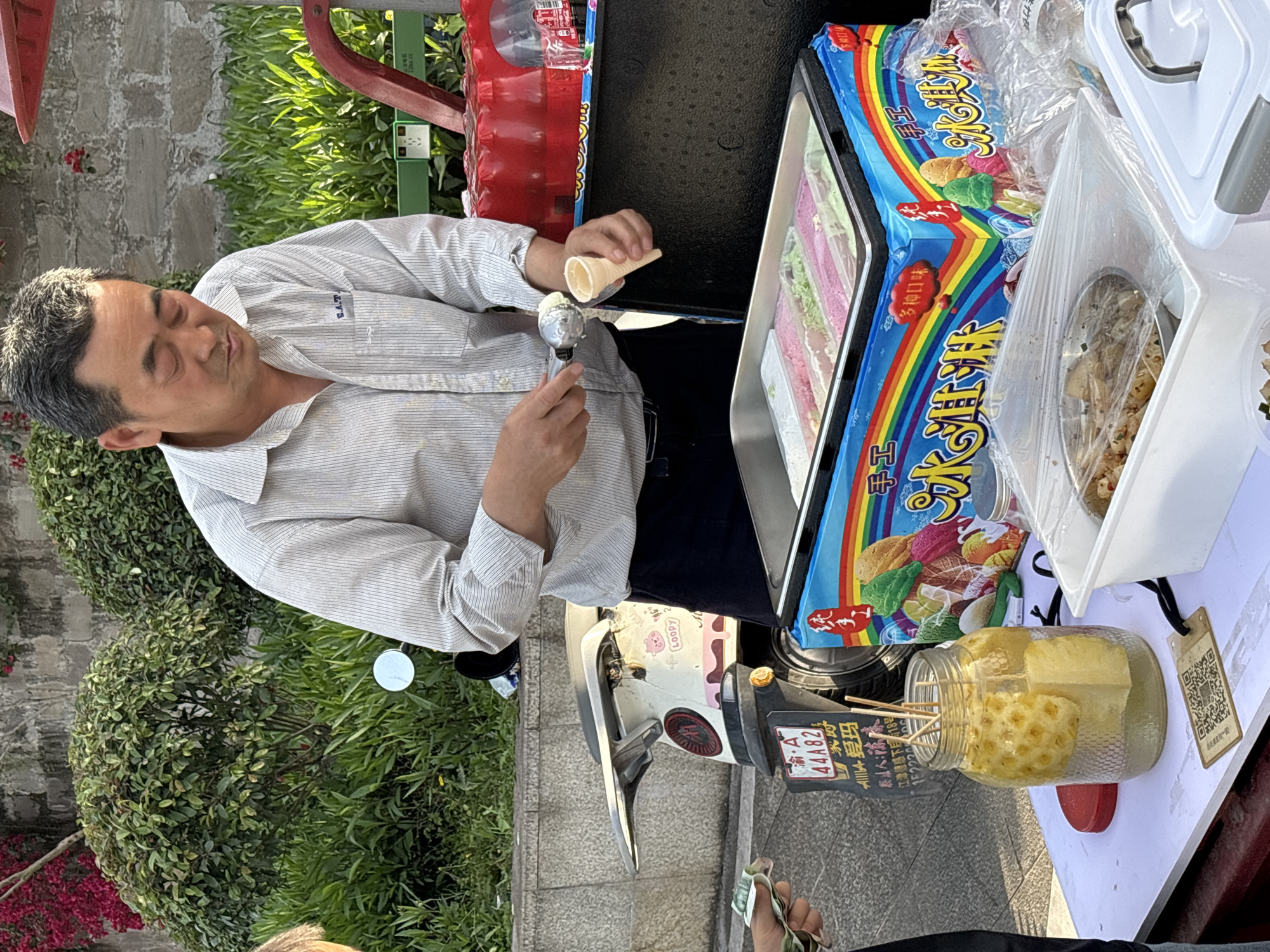 An outdoor scene depicts a male vendor preparing an ice cream cone from a mobile food stall. The man, with short, dark hair and grey streaks, is wearing a light-colored, striped collared shirt with "E.A.T." visible on the pocket, and dark trousers. He is focused, holding an ice cream scoop with a dollop of ice cream in one hand and an empty cone in the other.
His stall features a refrigerated display case containing various colored ice creams or sorbets (pink, green, white). Behind this, a large blue package with a rainbow design and Chinese text reads "手工" (handmade), "多种口味" (various flavors), and "手工冰淇淋" (handmade ice cream). Next to it are plastic containers, one with a white lid and another open, holding what appears to be a snack like popcorn. A glass jar filled with pineapple chunks on wooden skewers, submerged in liquid, sits on the counter, with a QR code attached to a tag on its side.
The vendor's setup is attached to a white scooter or moped, partially visible, which bears a license plate reading "A 44A82" and Chinese characters including "LOOPY" and "爱玛" (Aima, a brand). Further text on the scooter states "泰法人: 陈味" (Legal representative: Chen Wei) and "江津德感下月18号" (Jiangjin Degan next month 18th), along with a partial phone number "15222...". In the bottom right, a customer's hand holds banknotes, suggesting a transaction is underway. The background shows green bushes, a brick wall, and red plastic seating, all under bright daylight.