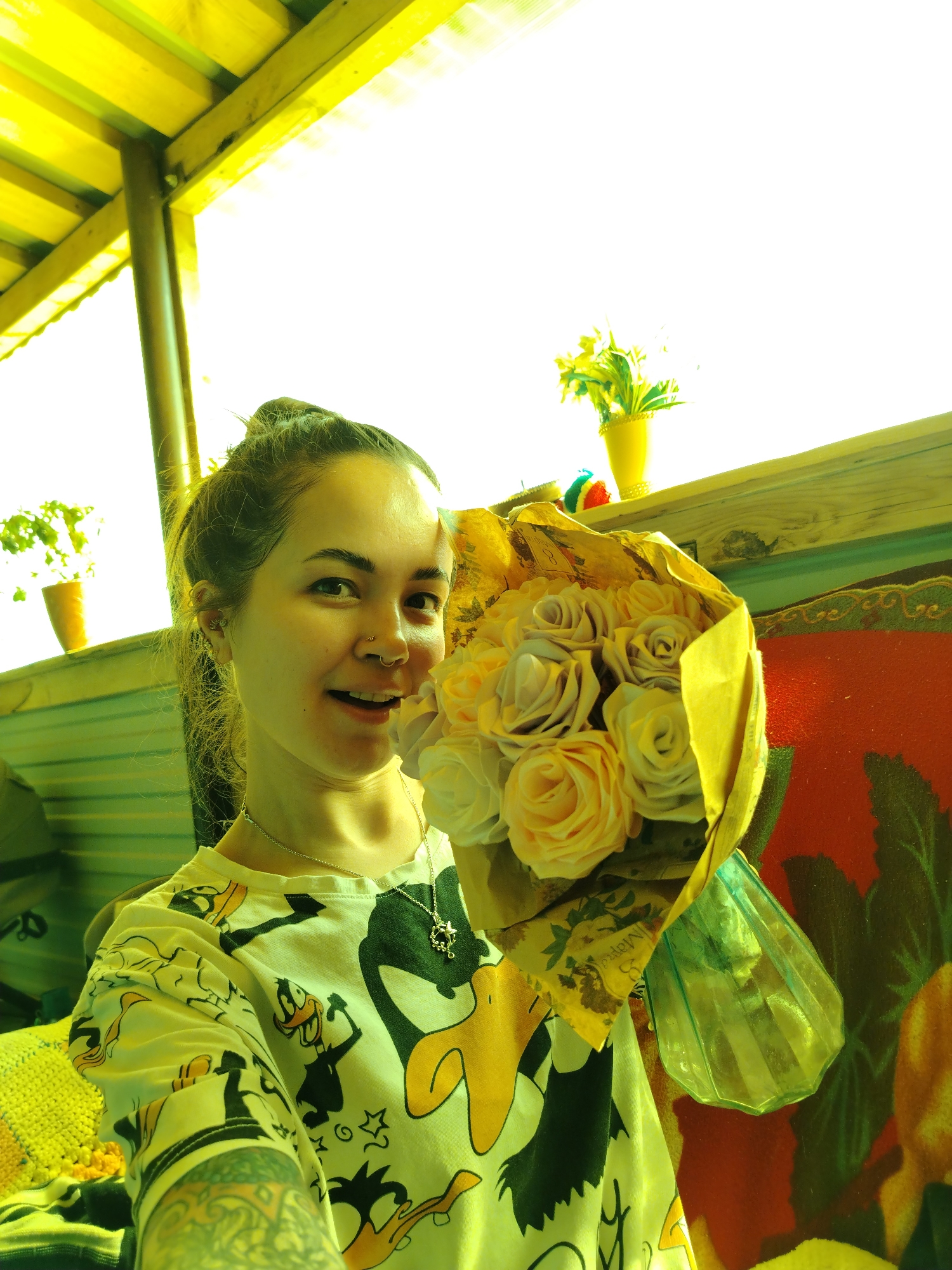 A woman is holding a bouquet of roses. She has her hair in a bun and is wearing a t-shirt with cartoon characters. She has a nose ring and a necklace. Behind her is a wooden structure, possibly a porch or deck, with a flowerpot on a ledge. A painting is behind the flowers. The location is Гожан, Russia.