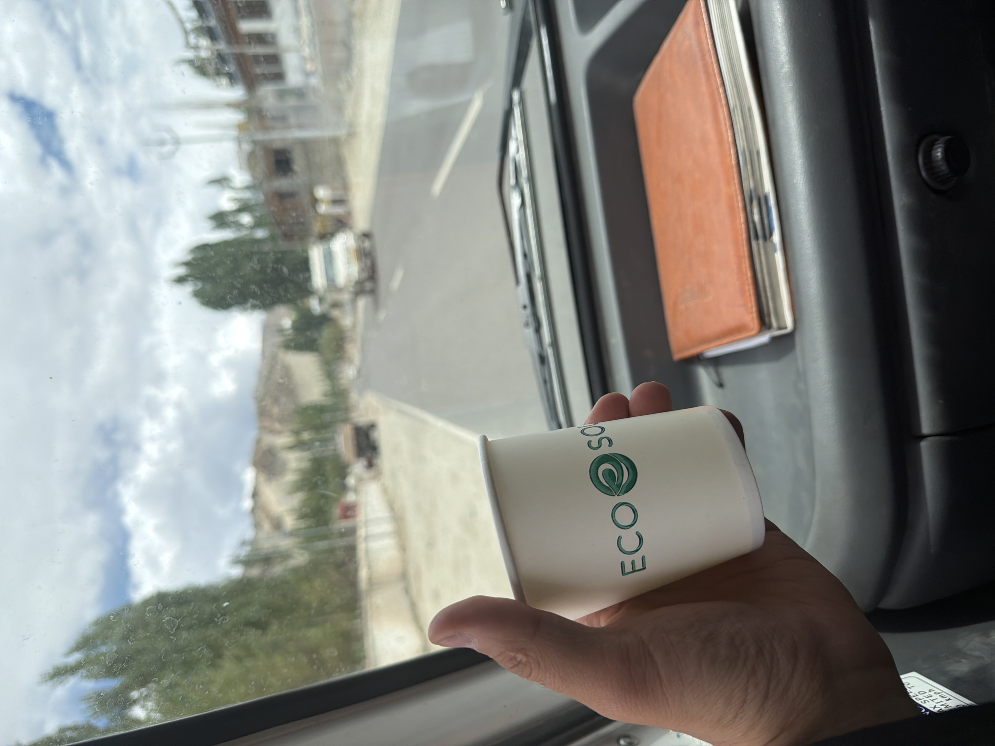 A hand holds a white paper cup with a green logo that reads "ECO SO". The cup is in the foreground, with a dashboard and a road scene visible through a vehicle's windshield in the background. The road is paved and appears to be in a somewhat rural or mountainous area, with buildings and trees lining its sides. The sky is cloudy. There is an orange notebook on the dashboard. The scene is likely in or near Thiksey, India, given the visual cues of the landscape and architecture.