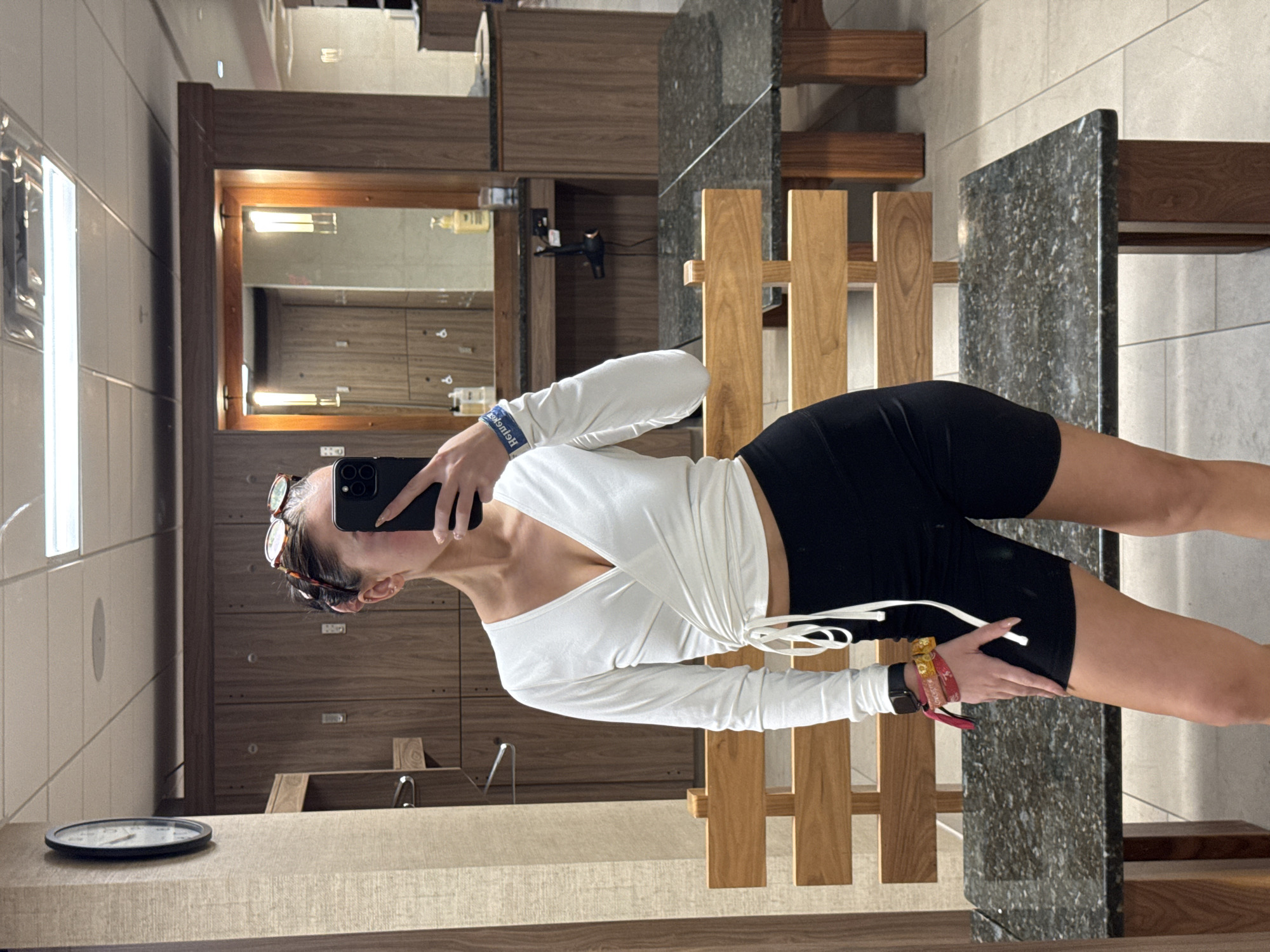 A woman is taking a mirror selfie in a restroom. She is wearing a white, long-sleeved wrap top and black biker shorts. She has a black phone with a dual camera held up to the mirror and a pair of sunglasses resting on her head. She is also wearing a watch and several bracelets. The restroom has brown wooden lockers, marble countertops, and wooden benches. The location is Irvine, United States of America.