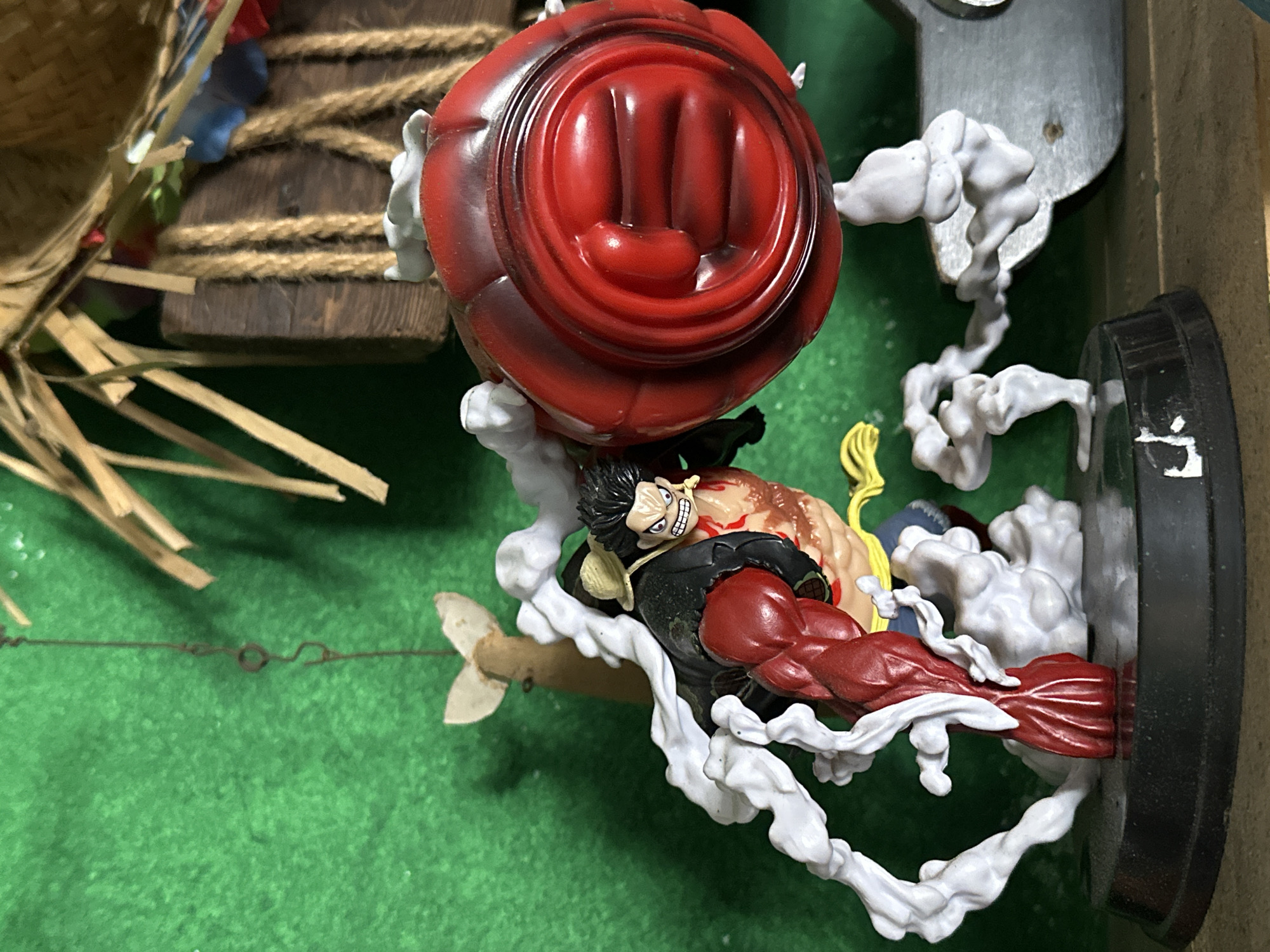 A detailed figurine of a character with red muscular arms, black hair, and a wide grin is lying amidst swirls of white cloud-like material on a green surface. Atop the figure is a large, red, rounded object shaped like a fist. A miniature thatched hut-like structure is visible in the background to the left. The figure is mounted on a black base, and a gray metal structure appears to the upper right. The scene is likely a display or a collection piece. The location in Andreyevka, Russia is not relevant to the description of the image itself.