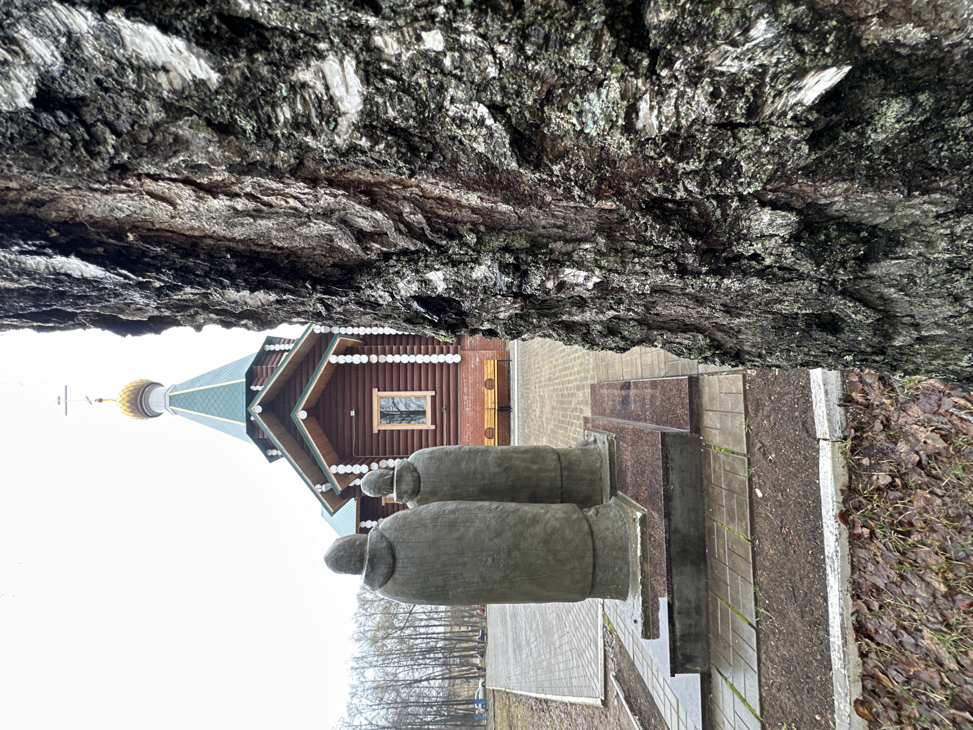 The image, viewed after being rotated 90 degrees clockwise, captures a serene religious scene in Chaikovsky, Russia, under an overcast sky. The immediate foreground is dominated by a large, textured section of dark tree bark, showing grey and green lichen, which appears to frame the upper portion of the view.
In the middle ground, a wooden chapel or church stands with a distinct architectural style. It is constructed from horizontal logs and features a green, multi-faceted pyramidal roof crowned by a golden onion dome and a small cross. White, spherical decorative elements adorn the eaves of the building, and a rectangular window is visible on its side.
Positioned in front of the chapel are two large, dark, monolithic statues on a paved platform. These abstract figures resemble hooded individuals or monks, crafted from stone or concrete, and stand side-by-side. Steps lead up to the platform where the statues are located.
The ground surrounding the structures consists of grey paver stones. To the right, patches of earth with fallen brown leaves and sparse grass are visible. In the background on the left, a line of bare deciduous trees suggests a colder season. The sky is uniformly white and overcast, indicating daytime on a cloudy day. No specific activity is depicted, and no text is visible within the frame, contributing to the quiet, contemplative atmosphere of the location.