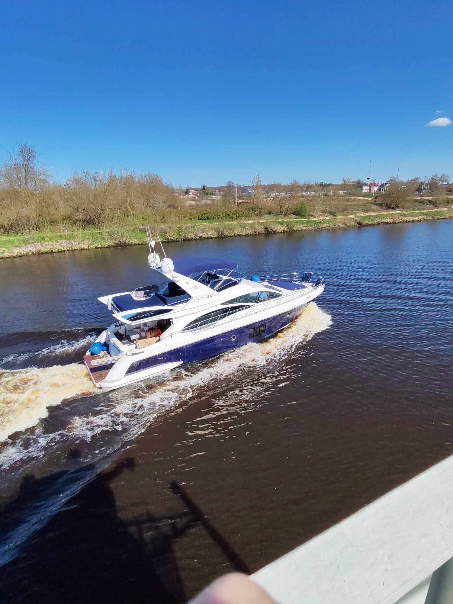 A medium shot captures a blue and white yacht speeding across a dark body of water, likely a river, leaving a trail of foamy wake behind it. The yacht is the primary focus, with its sleek design and visible upper deck. The river is flanked on both sides by grassy banks and trees, giving a natural setting. In the background, on one of the riverbanks, a small cluster of buildings can be seen, suggesting a nearby town or village. The sky above is a clear, vibrant blue, indicating a sunny day. The image is taken from a high vantage point, possibly a bridge, with a portion of the bridge's structure visible in the foreground. The absence of people suggests the yacht is either automated or the individuals onboard are out of view. The yacht's movement implies an action of transportation or leisure. The setting appears to be a rural or suburban area, with the town or village in the background adding a touch of human settlement to the scene. The overall atmosphere is serene and picturesque, with the dynamic movement of the yacht providing a contrast to the static landscape. The location is Unknown, Unknown.