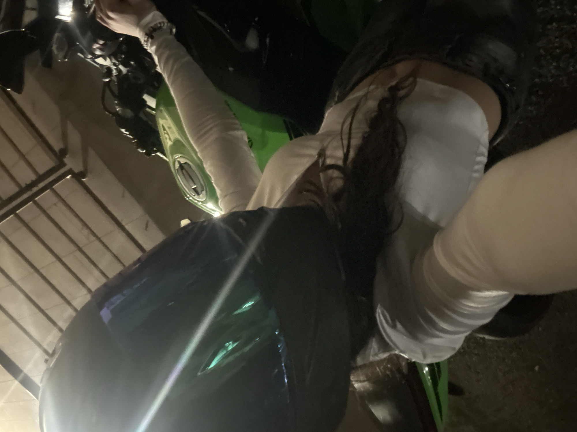 A person wearing a helmet is sitting on a Kawasaki Ninja motorcycle. The person has long dark hair, and is wearing a white long-sleeved shirt or jacket. The motorcycle is green and black. The image appears to have been taken at night. The environment suggests a garage or parking area under a roof with metal beams. There is no information that specifies the city or country. Yerevan, Armenia cannot be determined.