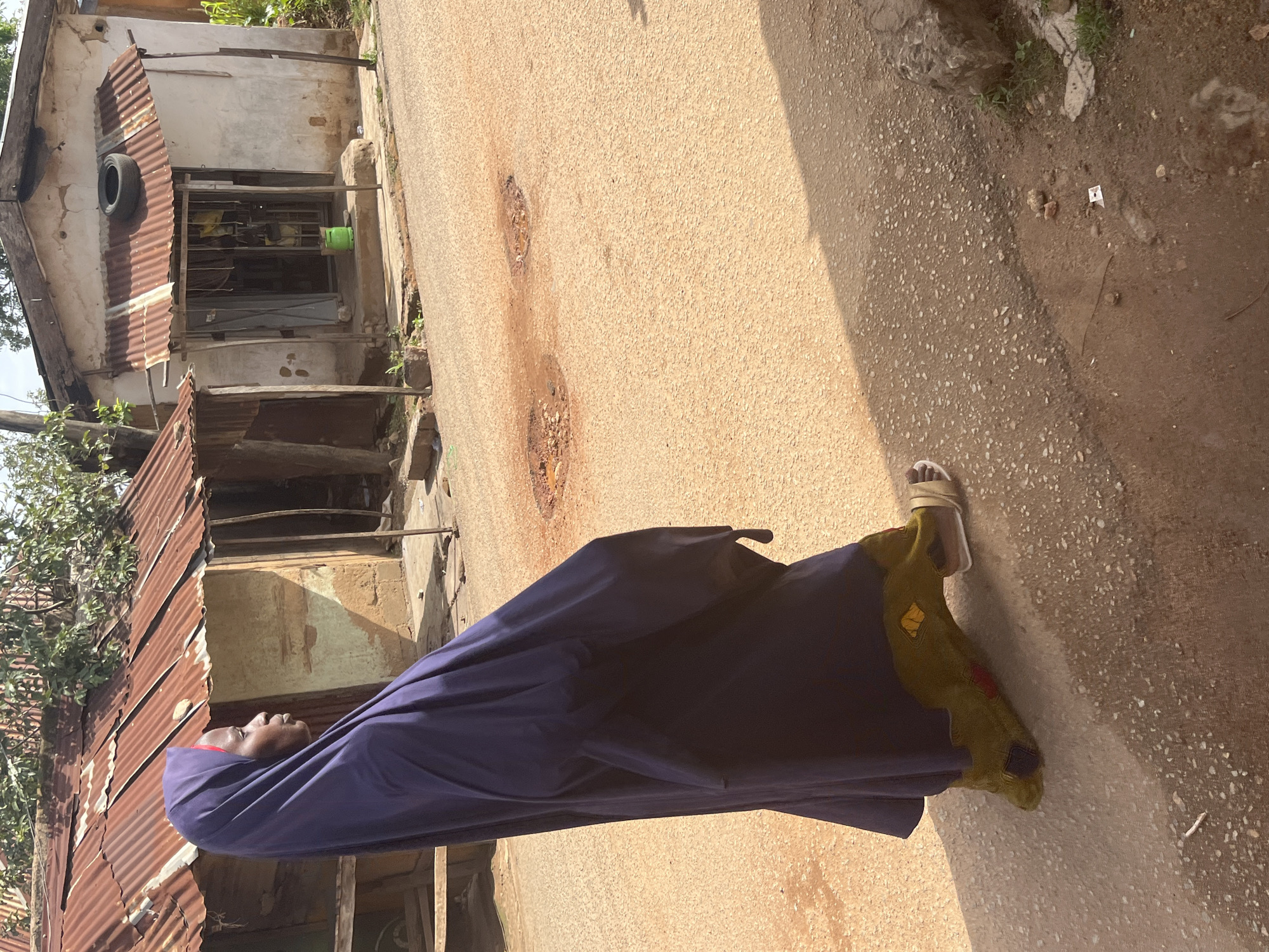 An individual in a dark purple hijab and a green and yellow patterned garment walks on a light brown dirt road in Jos, Nigeria. The individual wears light-colored sandals. To the left, a building with a rusty corrugated metal roof and mud walls is visible. To the right, another building with a white facade, a similar corrugated metal awning, and a dark opening is present. The road has several patches of standing water and exposed soil, suggesting recent rain. The scene is illuminated by bright daylight.