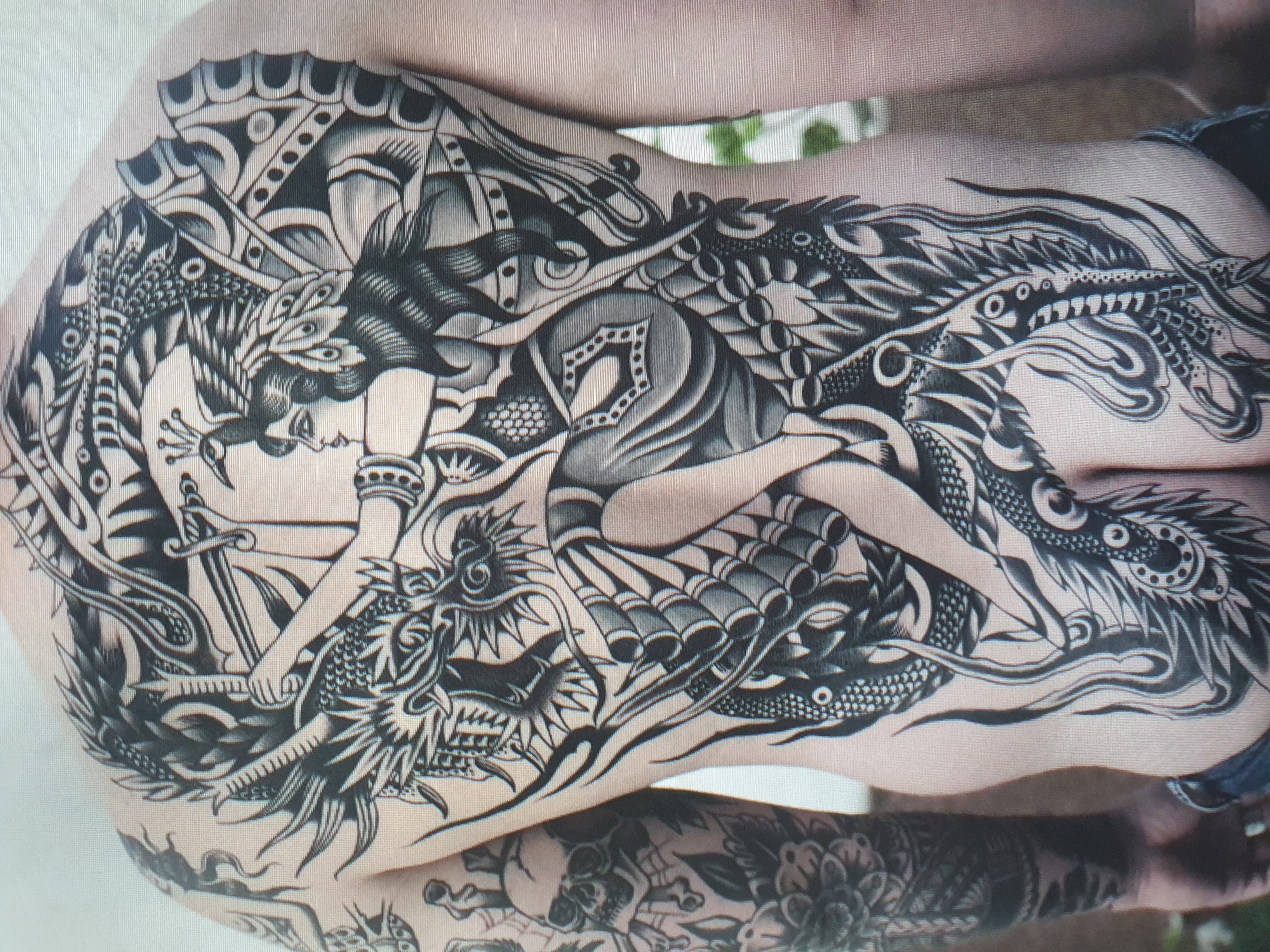 The image shows a person with a large black and grey tattoo covering their back and extending down their side. The tattoo depicts a dragon entwined with a female figure holding a sword. The style appears to be traditional Japanese or American traditional tattoo art. There are also elements of a skull and floral motifs visible on the person's arm. The background is out of focus, but appears to be an indoor setting. There is no information in the image to suggest a location in Tashkent, Uzbekistan.