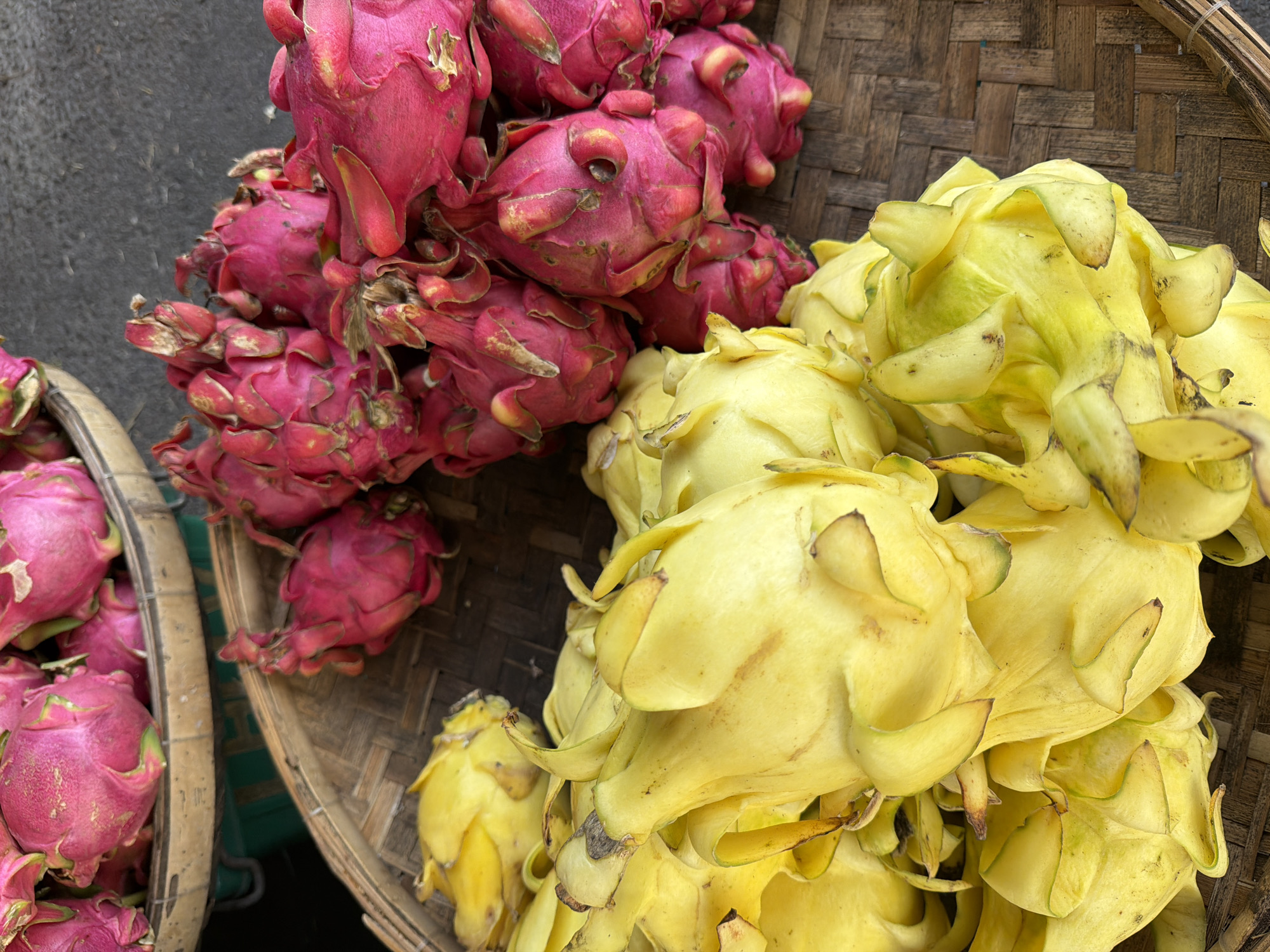 In the city of Nha Trang, Ward, Vietnam, a close-up, high-angle shot captures a collection of vibrant dragon fruits. In the foreground, a dense pile of bright yellow dragon fruits fills the majority of the frame, their distinctive scales and leafy protrusions clearly visible. Behind the yellow fruits, and slightly to the right, a pile of intensely pink dragon fruits is present. These pink fruits are also piled high, with their characteristic winged scales dominating their appearance. Both piles of fruit are resting on woven bamboo trays, which create a textured, earthy background for the colorful produce. The overall lighting suggests an outdoor setting, possibly a market stall.