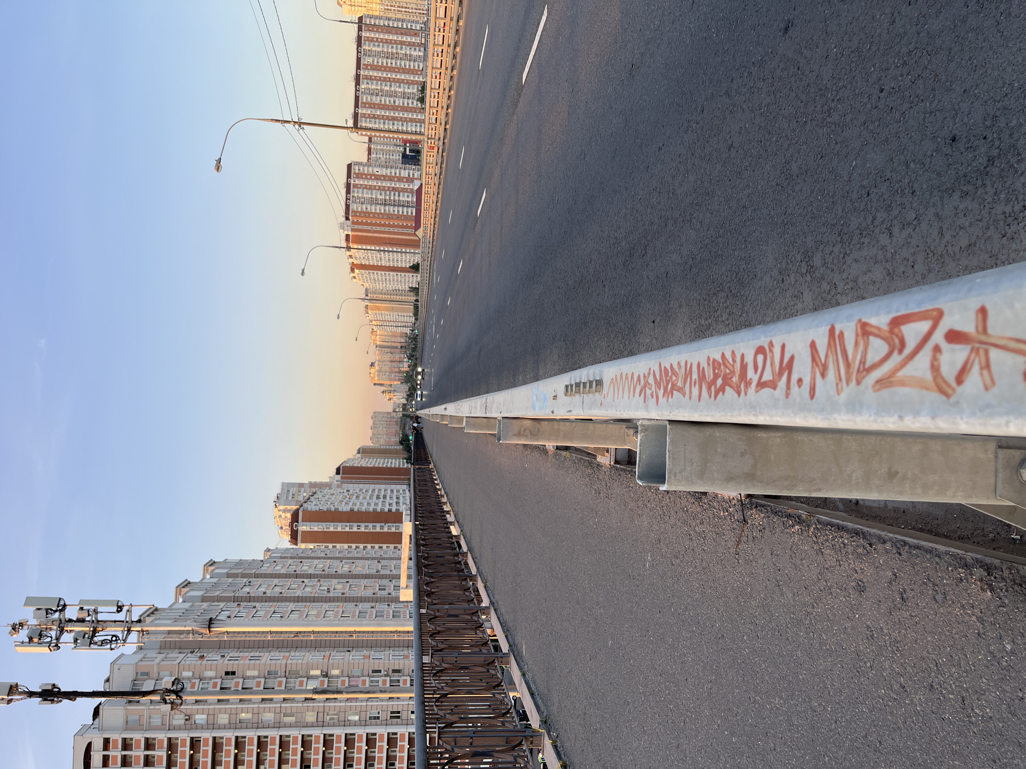 The image depicts a view from a bridge in Lyubertsy, Russia, looking down a multi-lane road towards a cityscape of residential buildings under a clear, pale sky. On the left side of the road is a pedestrian walkway bordered by an ornate metal railing. The road itself is asphalt, with white lane markings. Several streetlights are visible along the roadside. In the distance, a dense cluster of tall, modern apartment buildings, primarily with light-colored facades and some red accents, dominates the horizon. The buildings have numerous windows and balconies, indicative of a residential area. Some buildings feature antennas or communication towers on their roofs. The lighting suggests it is either early morning or late afternoon, with a soft, warm light casting long shadows. Graffiti in red spray paint is visible on a metal barrier in the foreground. The scene is devoid of people but shows signs of vehicular traffic in the distance.