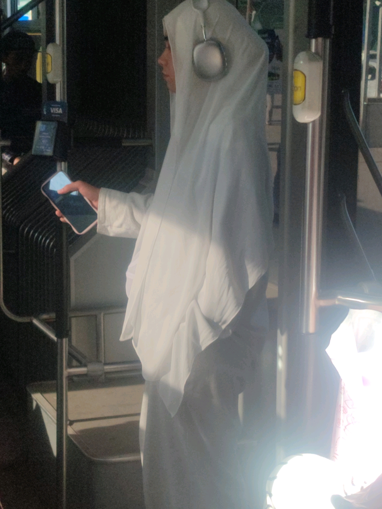 A young woman wearing a white hijab and headphones is standing inside a bus, holding a smartphone in her right hand. She is looking to her right, away from the camera. The bus interior is visible, with metal poles, seats, and a ticket validator. Sunlight is streaming in through the windows, creating strong highlights and shadows. In the background, another person is visible in silhouette. The environment appears to be a public transport setting.