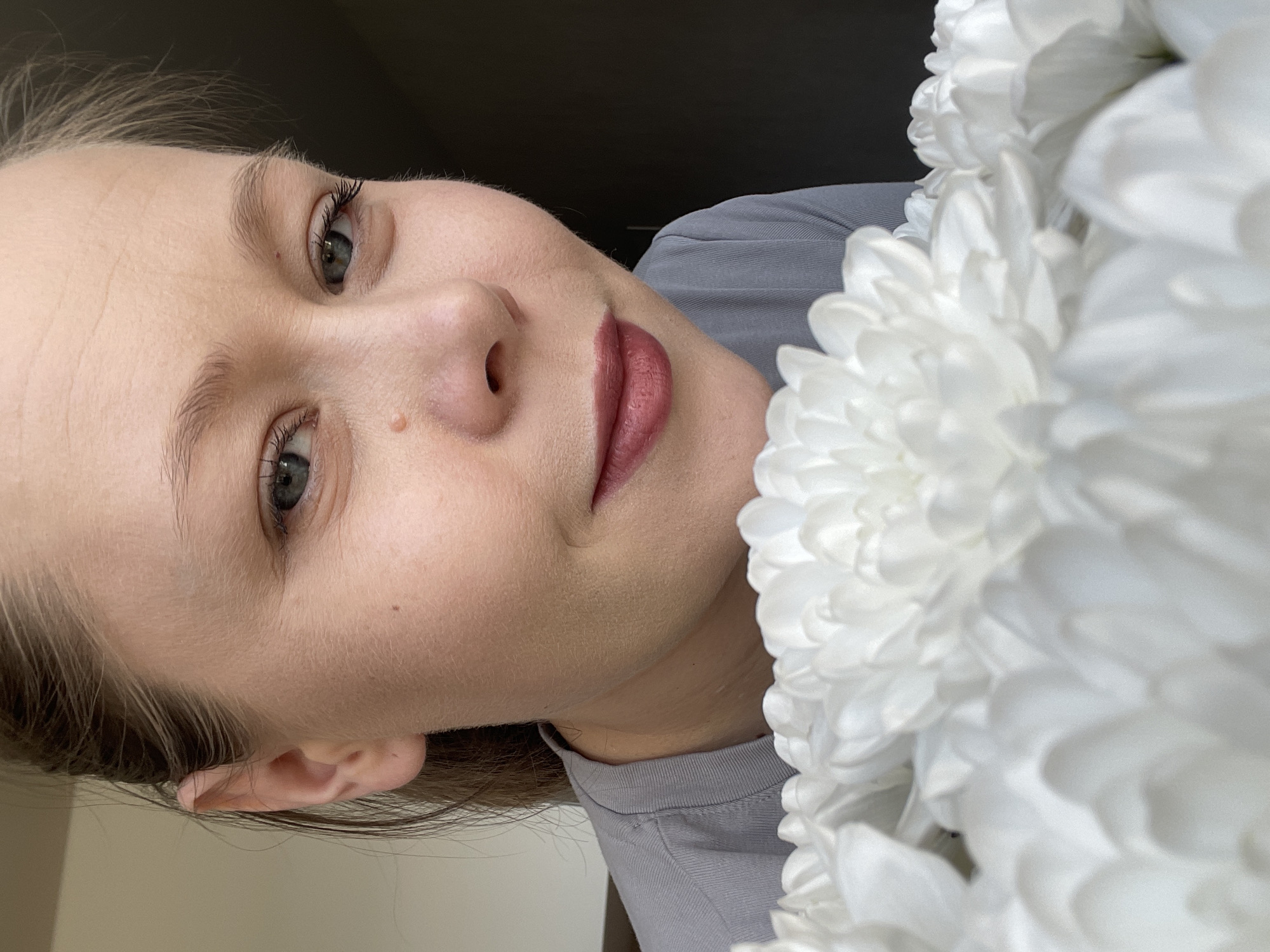 This media file features a close-up, tilted portrait of a young adult woman indoors, presenting a calm expression. She has fair skin, with light brown hair pulled back, visible on the sides. Her green or grey eyes are accented by long, dark eyelashes and defined eyebrows. A small mole is visible on her left cheek and another on her forehead. Her lips are full and tinted with a pinkish-red lipstick. She is wearing a plain grey t-shirt.
To her right, a substantial bouquet of white, fluffy flowers, likely chrysanthemums, dominates the frame, with some petals in the foreground appearing slightly out of focus. The background behind the woman's head is dark and plain, contrasting with a lighter, plain surface on her left. The lighting is soft and even, suggesting an indoor setting, possibly with diffused natural light. No visible text is present in the image. The location context provided for this image is Chaikovsky, Russia.