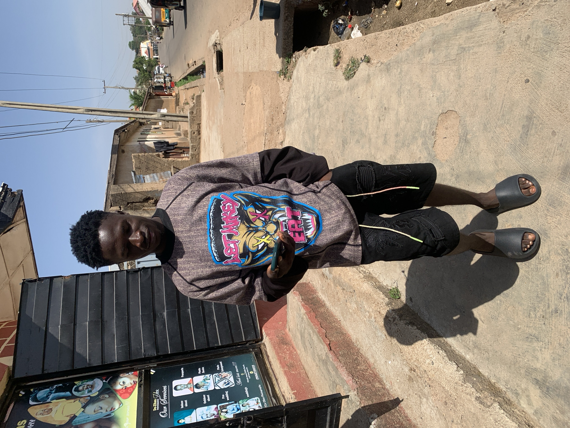 A young man stands in front of what appears to be a business establishment with a black corrugated metal exterior in Jos, Nigeria. The street view features buildings and utility poles under a bright sky. The man is wearing a patterned shirt and black shorts, and slides. He is holding a smartphone. A poster is visible on the front of the building, displaying text about photography services. The pavement shows wear and tear.
