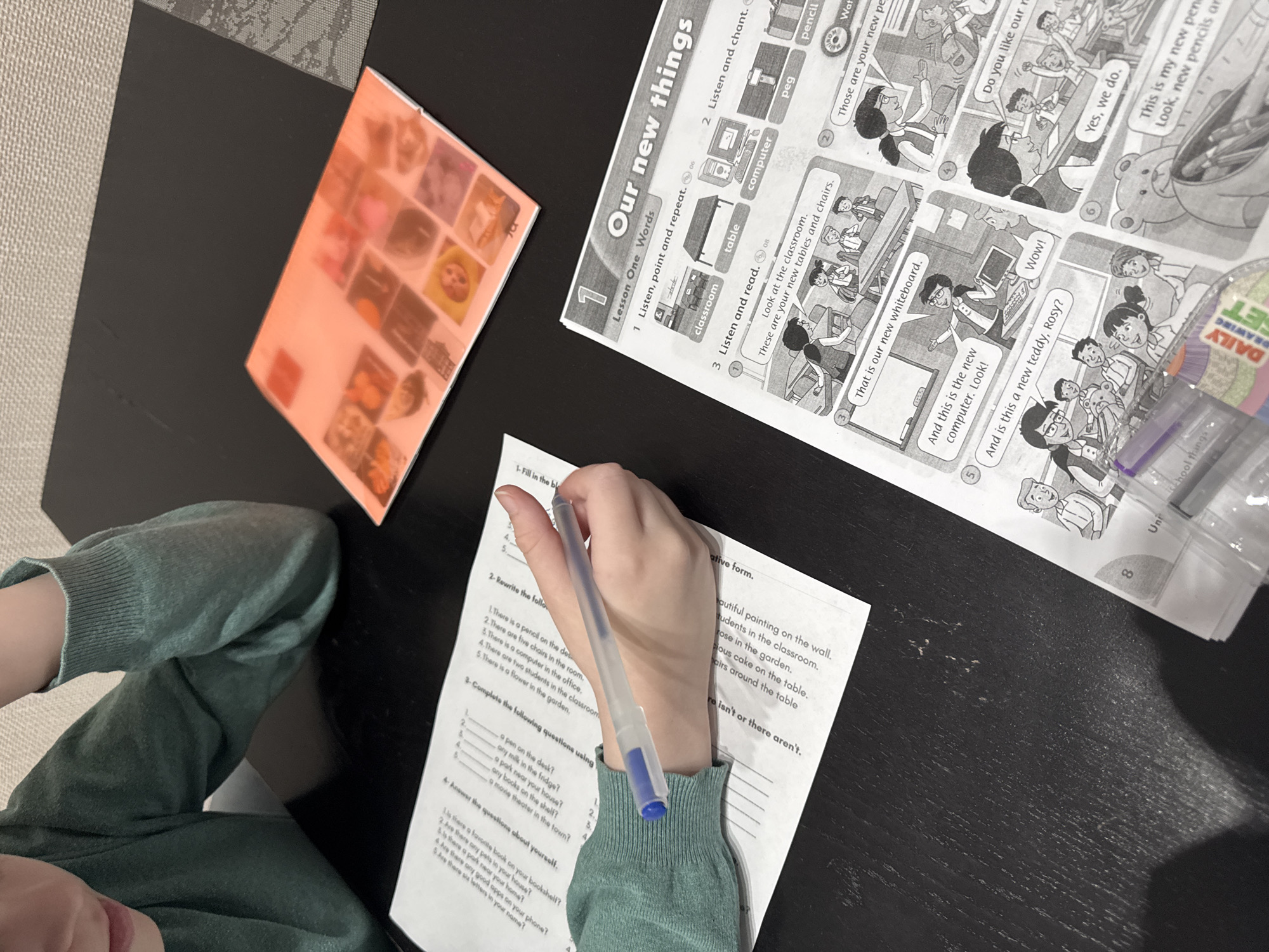 A child is completing a school worksheet on a dark wooden table. The worksheet focuses on forming questions and negative sentences in English related to classroom and home environments. The child is writing with a blue pen. The textbook, "Our New Things", is open to a page showing a classroom scene with new items like a whiteboard, computer, and teddy bear. Next to the textbook, there is an orange book cover. A child's arm and leg are visible, wearing a light green sweater. The location and city are not discernible from the image.