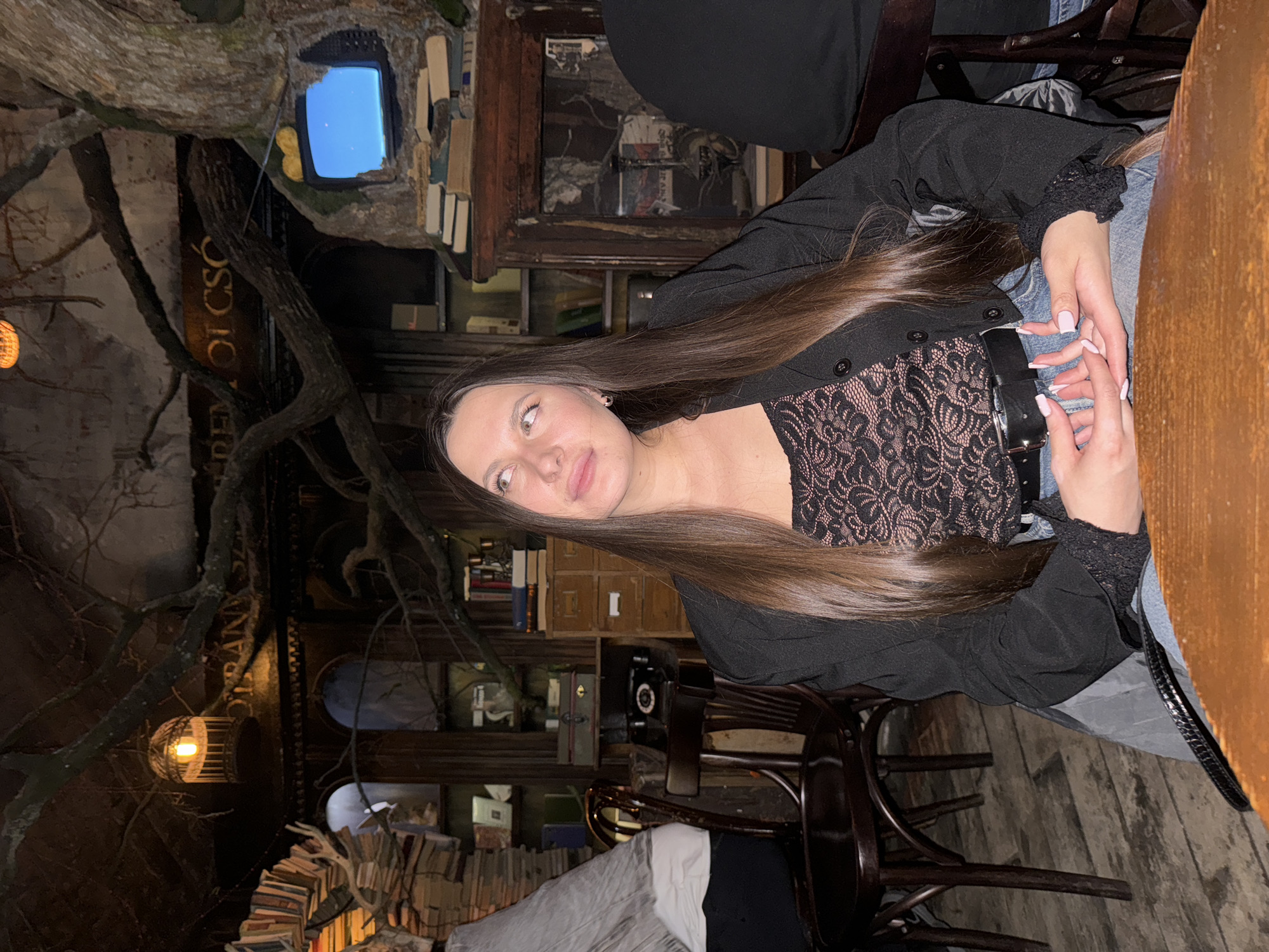 A young woman with long brown hair sits at a wooden table in what appears to be a uniquely decorated restaurant. She is wearing a black blazer over a black lace top, with jeans and a black belt. Her hands are clasped together on the table. The restaurant has a distinctive interior, with branches and decorative elements that look like a tree growing inside. Shelves of books line the walls. In the background, there is a retro television set embedded in the decorative tree structure. The specific location is the restaurant "Odessia" in Moscow, Russia.
