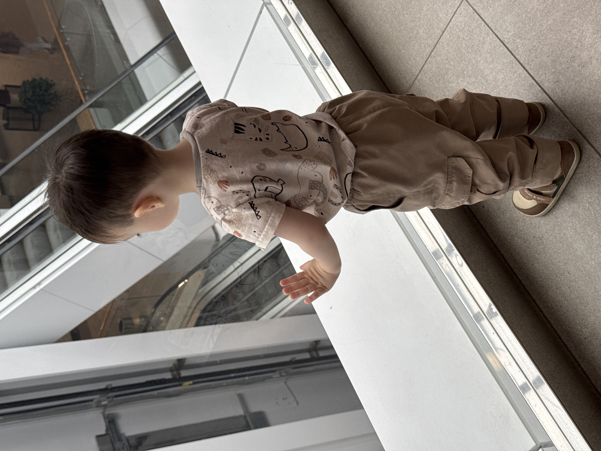 A child is standing near a large window overlooking a building with escalators. The child is wearing a beige shirt with animal patterns and beige pants. The child's shoes are brown with green trim. The child is looking out the window. The location cannot be determined from the image.