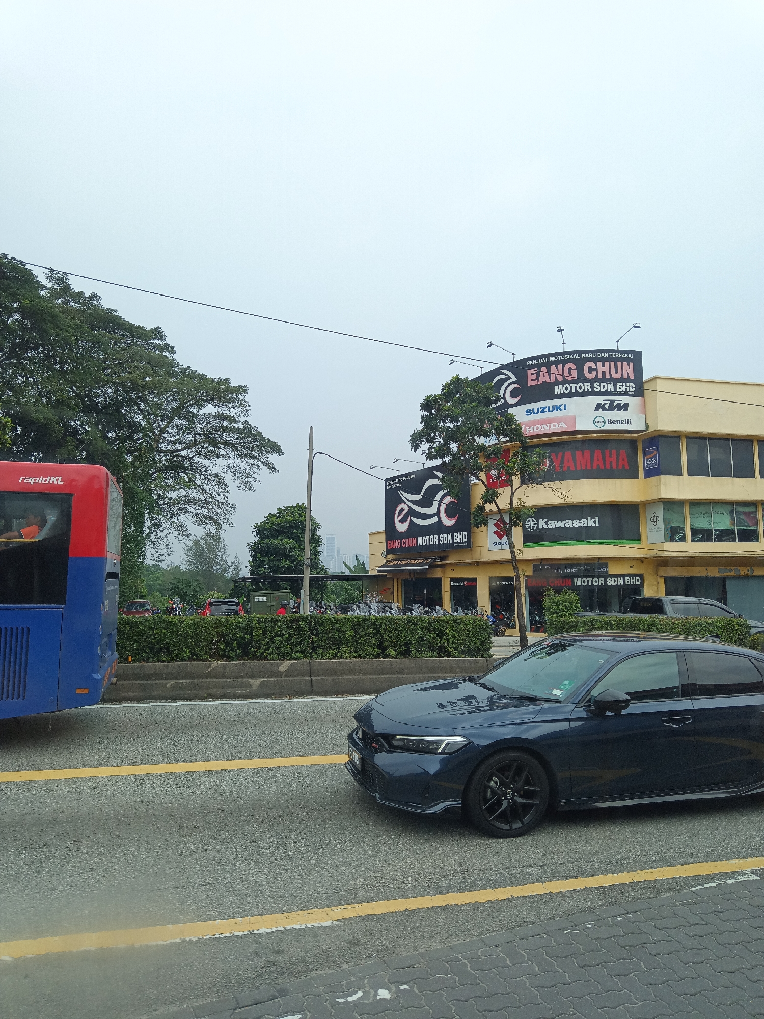 A dark blue sedan is parked on the right side of the road in Kuala Lumpur, Malaysia, during the daytime. The car is facing towards the right of the frame, with its front wheels turned slightly. In the background, a yellow building with the signage "EANG CHUN MOTOR SDN BHD" is visible, along with various motorcycle brand logos such as Yamaha, Kawasaki, Suzuki, Honda, Benelli, and KTM. A red and blue bus with "rapidKL" branding is on the left side of the road, moving away from the camera. The sky is overcast, suggesting a cloudy day. Trees line the roadside, and a pedestrian walkway with patterned paving stones is present in the foreground.
