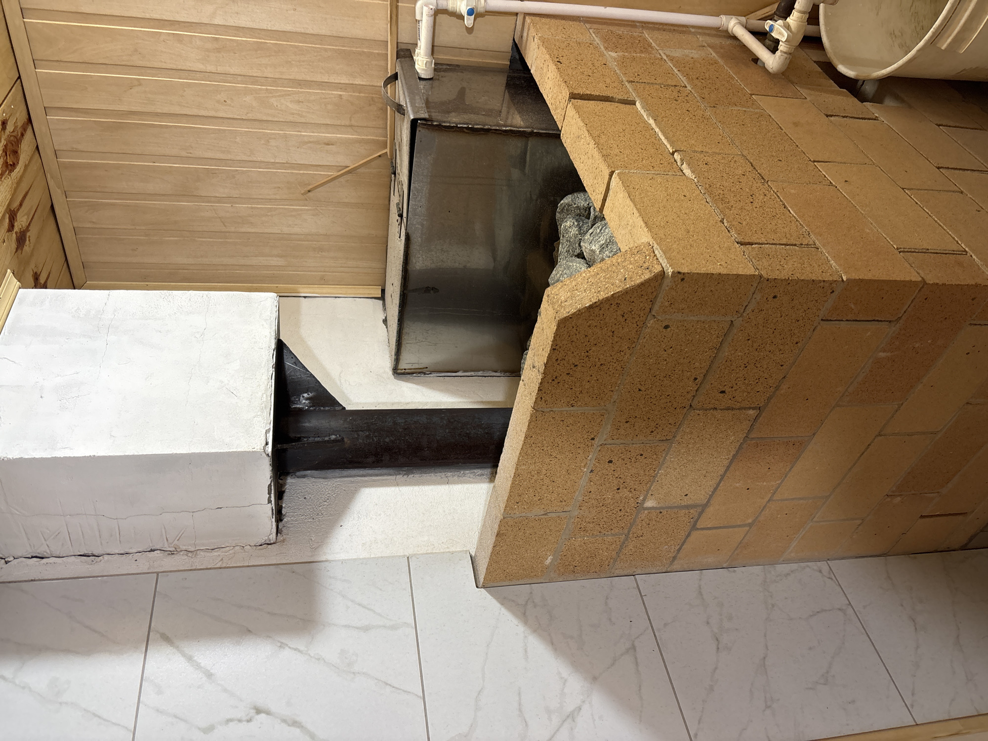 In the city of Ilpanur, Russia, a Finnish sauna is pictured. The focal point is a brick oven with a metal enclosure filled with rocks, likely for heating. To the left, a wall is clad in white tiles with a marbled pattern. To the right and overhead, the wall and ceiling are constructed of light-colored wood planks. A metal pipe protrudes from the oven and extends upwards, surrounded by a white, squared structure. Adjacent to the oven, plumbing fixtures, including a pipe and valve, are visible, along with a white bucket situated on the floor.
