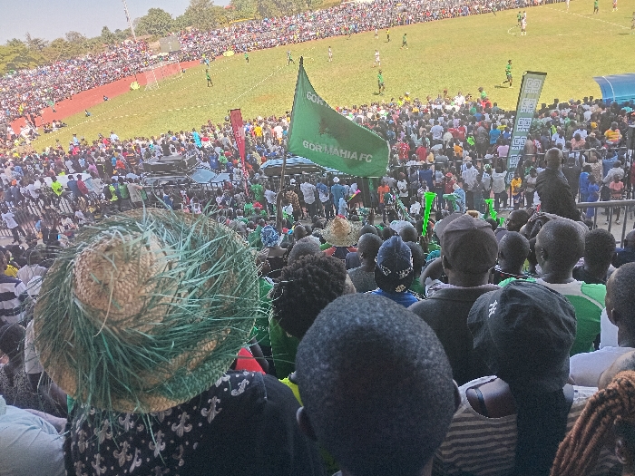 This photograph captures a bustling football stadium in Siaya, Kenya, during a live match. The perspective is from within the spectator stands, looking down towards the pitch and across a densely packed crowd.
The stadium is filled with numerous spectators, many of whom are seen from behind or as a sea of figures in the distance. In the immediate foreground, the backs of several heads are visible, including one individual wearing a distinctive straw hat adorned with green, straw-like material. The crowd extends far into the background, suggesting a well-attended event.
A large, green flag prominently displays the text "GOR MAHIA FC," identifying one of the competing teams or the favored club of the fans. Many spectators appear to be wearing green, further indicating support for Gor Mahia FC. Along the perimeter of the football pitch, various advertising banners, including one with a "GG" logo, are visible.
On the green playing field, football players in green and white kits are actively engaged in the match. Goalposts are visible at both ends of the pitch. The scene is set under clear, bright skies, suggesting good weather conditions for the outdoor event, likely during daylight hours. The vibrant crowd and prominent team flag convey an atmosphere of excitement and strong team support.